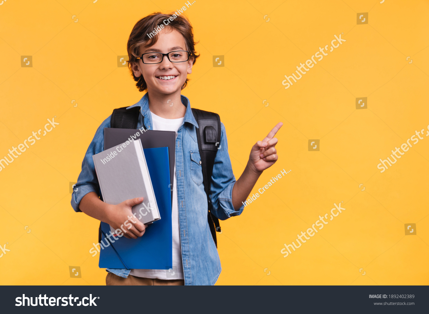 Smiling little boy pointing at copy space in casual clothes with books for studing at school isolated over yellow background #1892402389