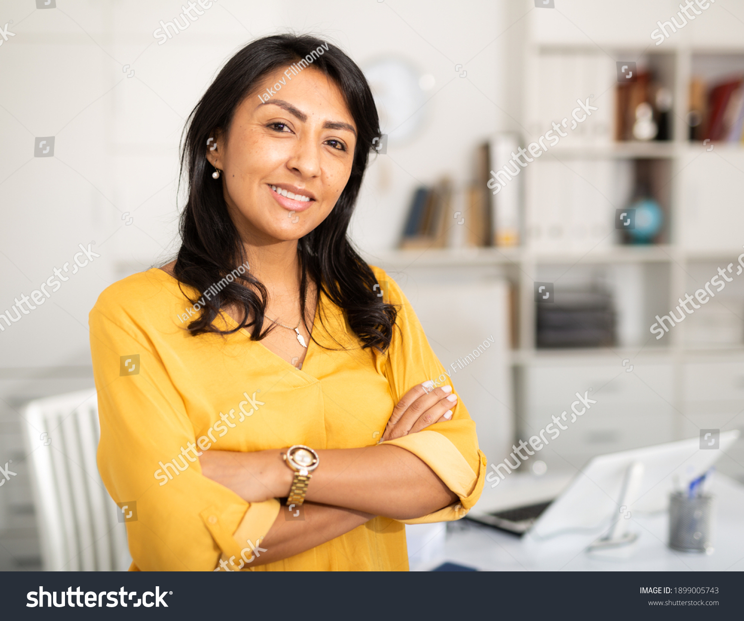Happy smiling Latin American businesswoman standing in office with arms crossed looking at camera #1899005743
