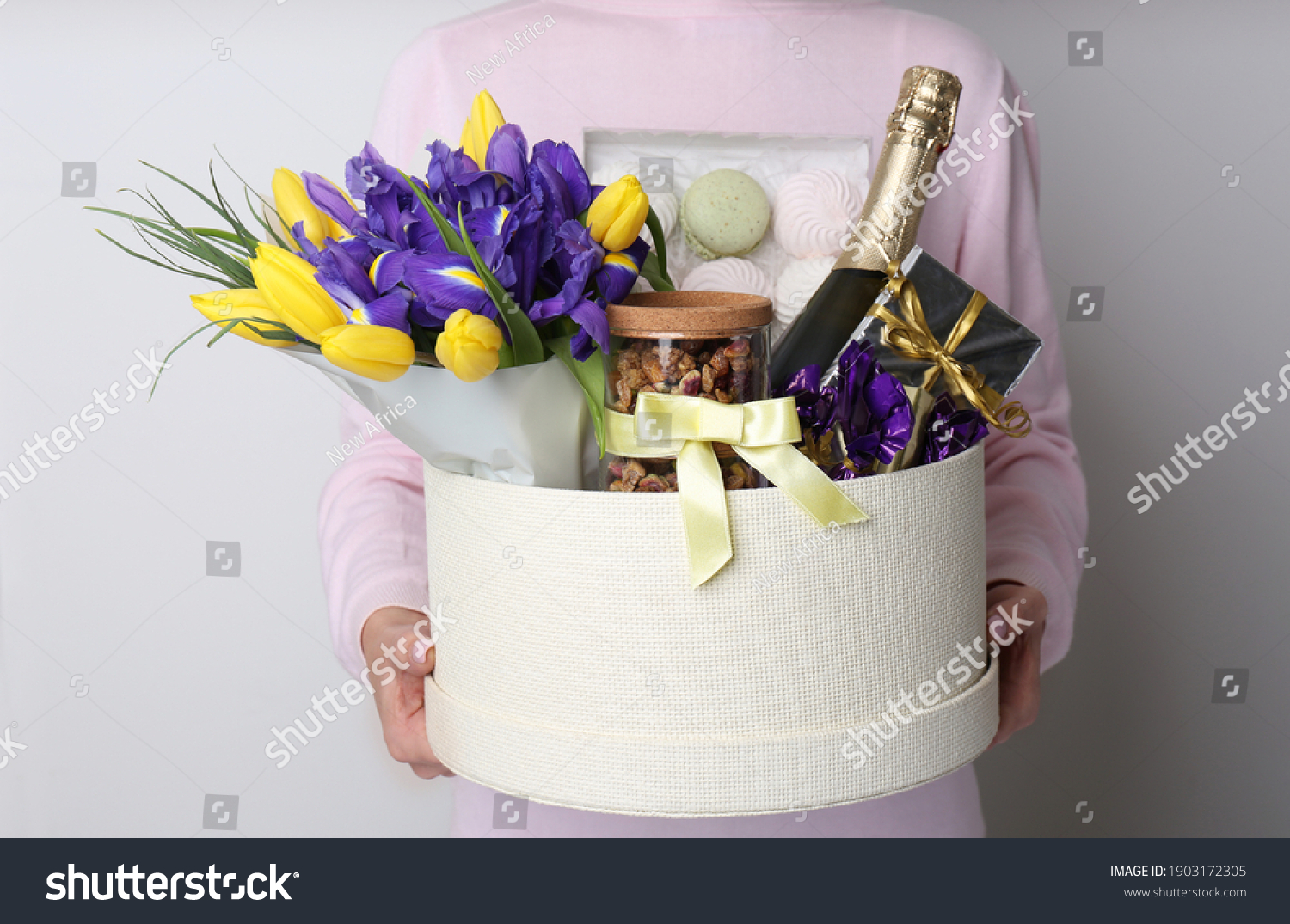 Woman holding basket with gifts, bouquet and champagne on light grey background, closeup #1903172305