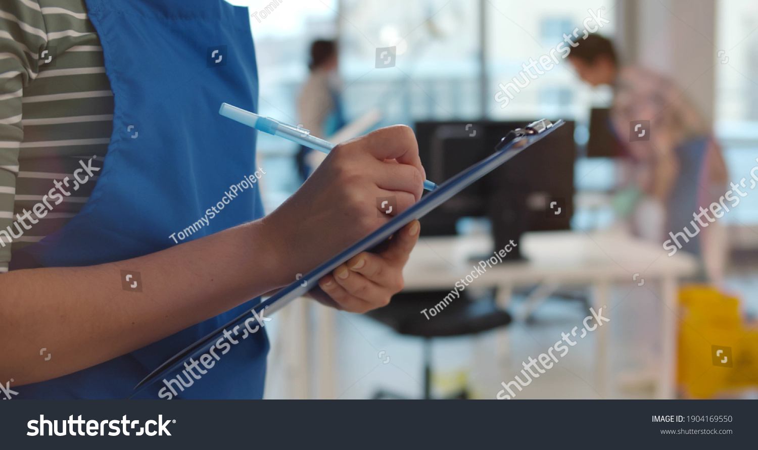 Team of professional janitors in uniform cleaning office. Close up of woman cleaner checking list on clipboard supervising cleaning service team work in modern office #1904169550