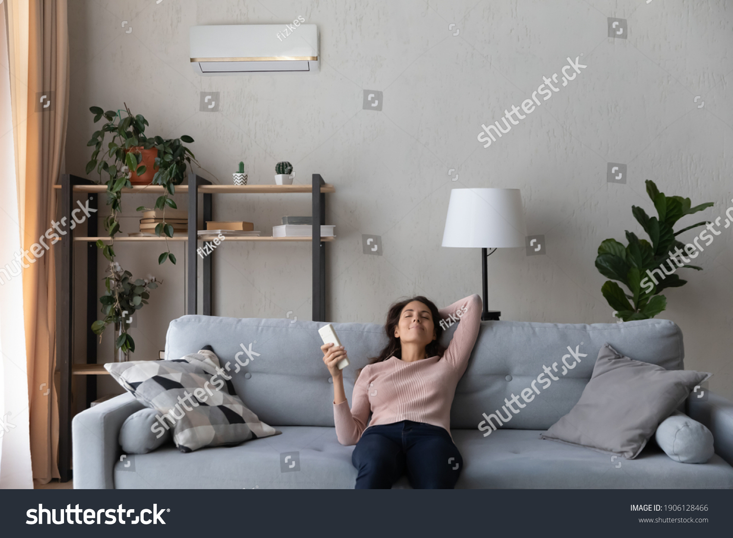 Happy young caucasian woman turning on air conditioner with controller, breathing fresh cooled air or enjoying comfortable temperature indoors, relaxing on comfortable sofa in modern living room. #1906128466