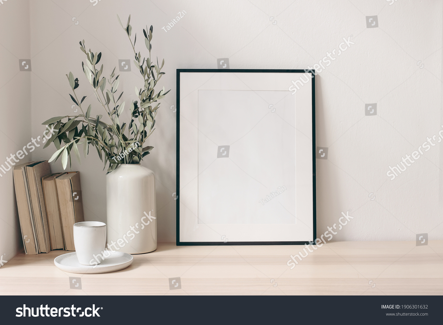Breakfast still life. Cup of coffee, books and empty picture frame mockup on wooden desk, table. Vase with olive branches. Elegant working space, home office concept. Scandinavian interior design. #1906301632