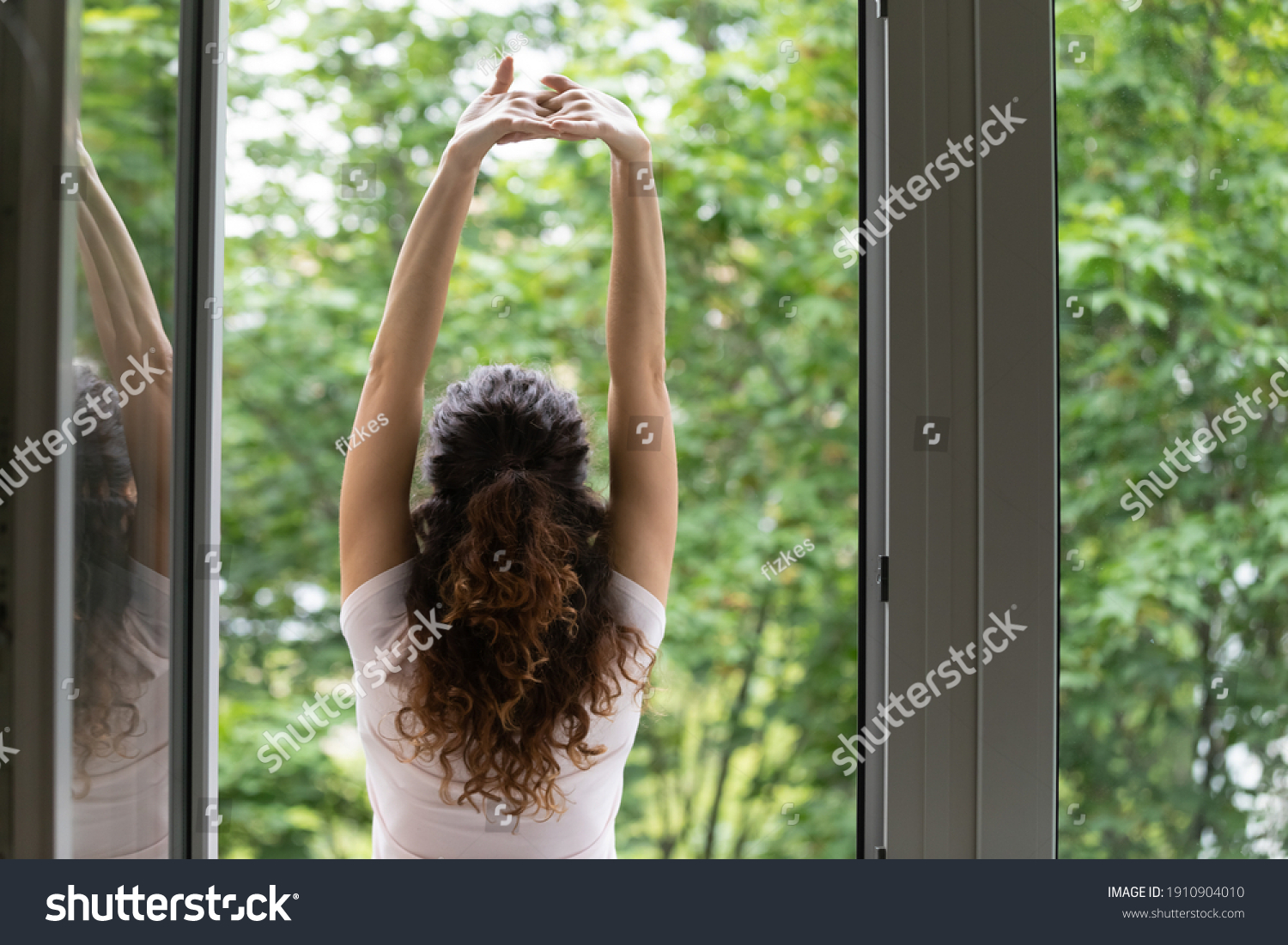Meeting summer day. Back rear view of happy young woman stand in balcony door stretch hands enjoy weekend have pleasure. Calm beautiful female do morning exercises practice yoga by opened large window #1910904010