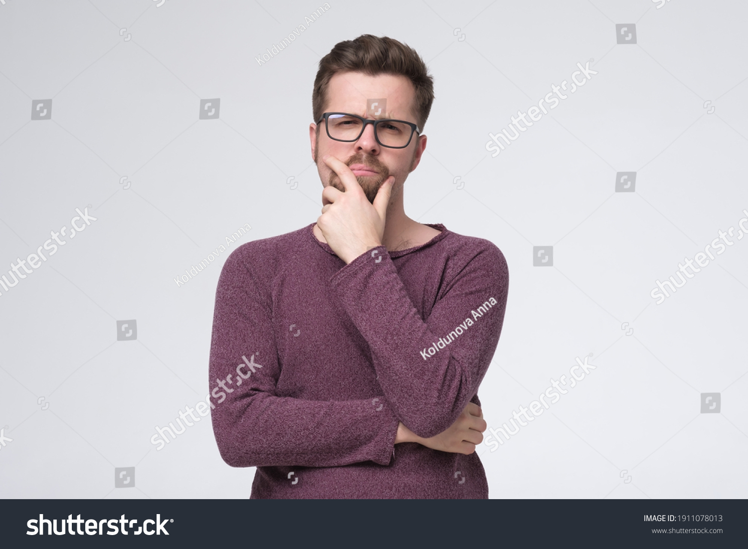 Generating new ideas for you. Thoughtful young man holding hand on chin and looking at camera while standing isolated on white wall #1911078013