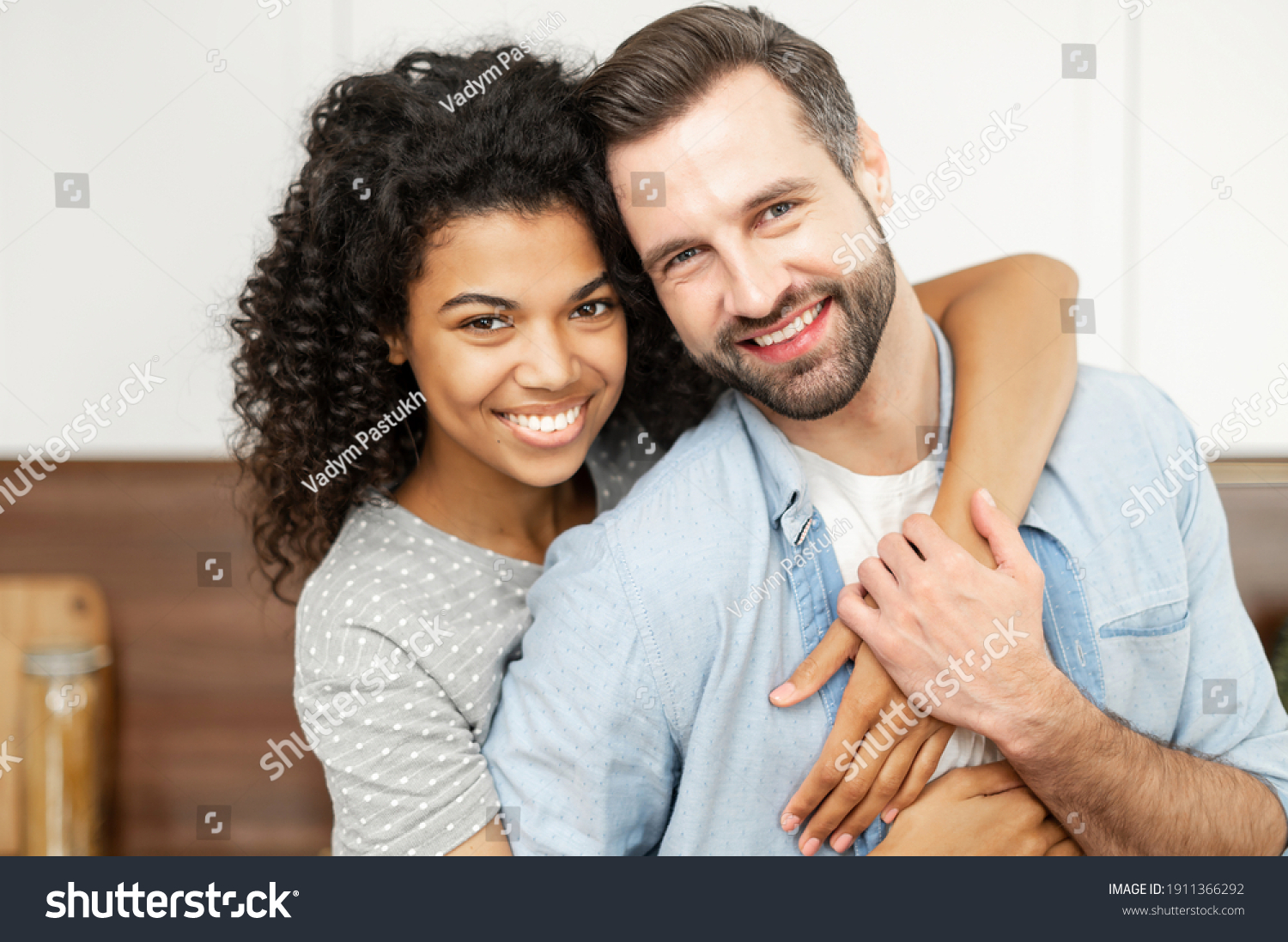 Close-up portrait of happy multi-ethnic couple in love. An African woman hugs a caucasian guy from behind, they looks at the camera with a cheerful smiles #1911366292