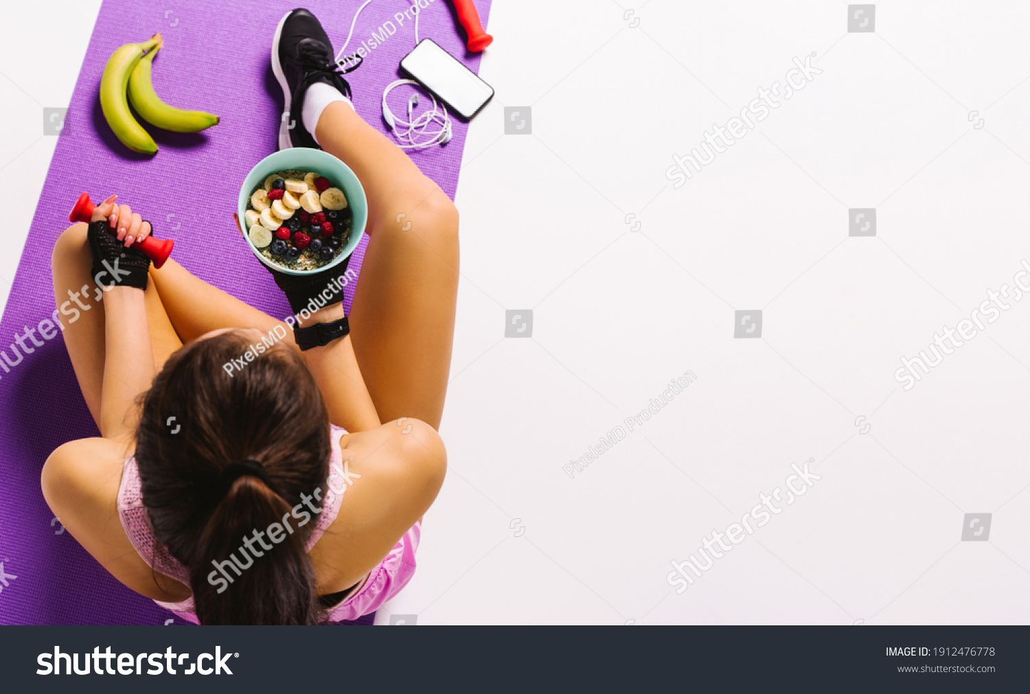 Sports and nutrition. Top photo of a girl with dumbbells and muesli in hand while sitting on the mat. White background and blank advertising space. #1912476778