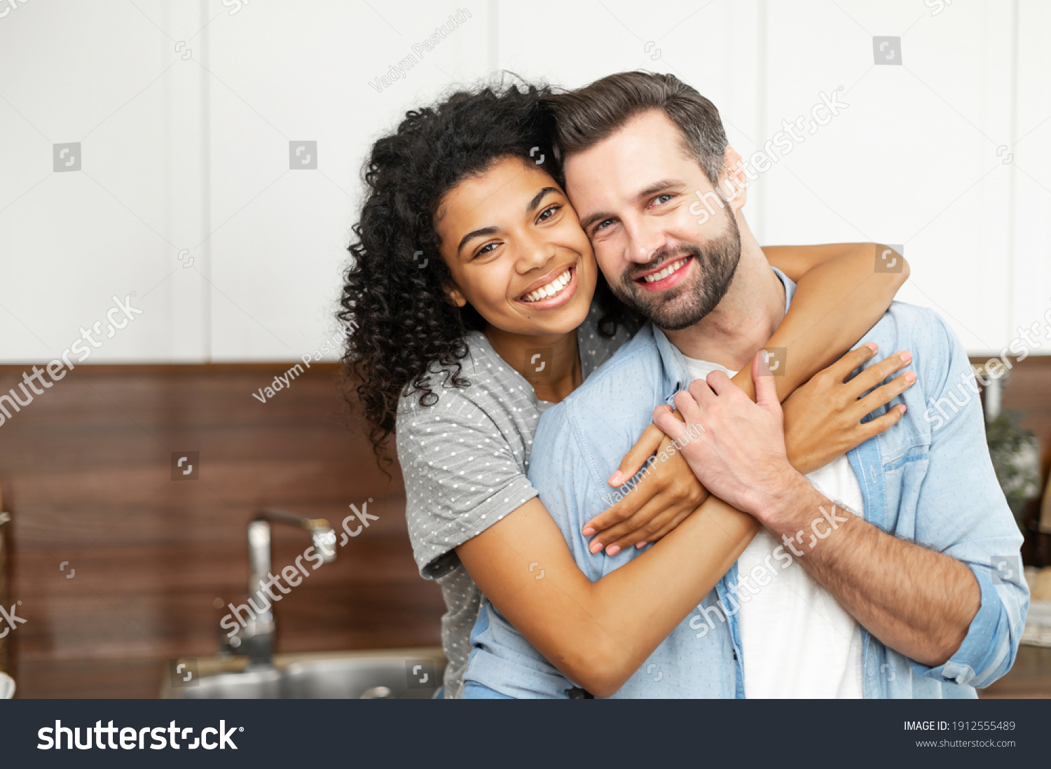 Close-up of happy interracial couple posing over blurred kitchen background, happy owners of new flat smiling and looking at the camera, young African American woman hugging handsome man from behind #1912555489