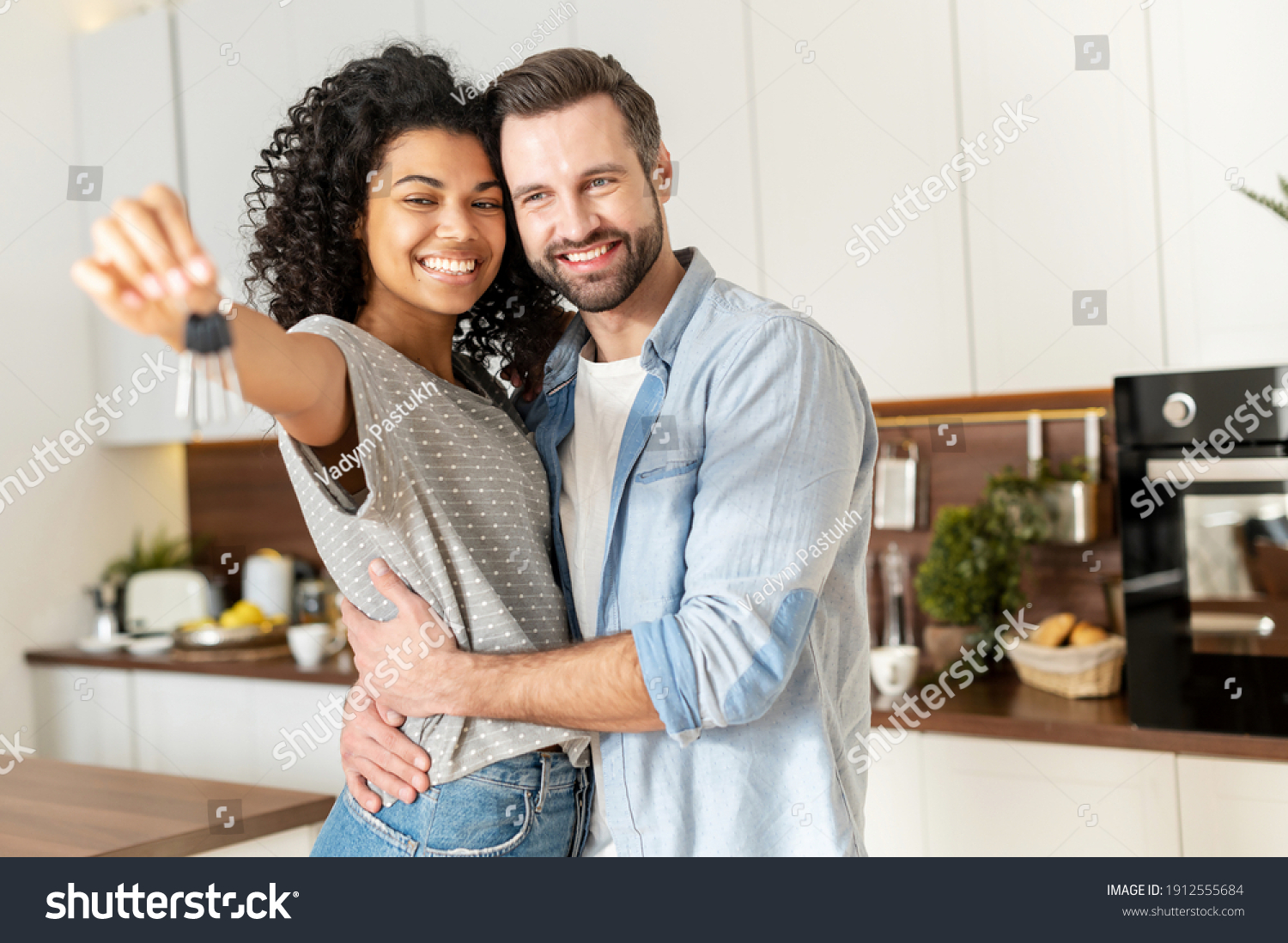 Young interracial married couple homeowners smiling, showing keys from a new apartment, hugging and looking at the camera, standing in the kitchen and celebrating moving in a new home, family concept #1912555684