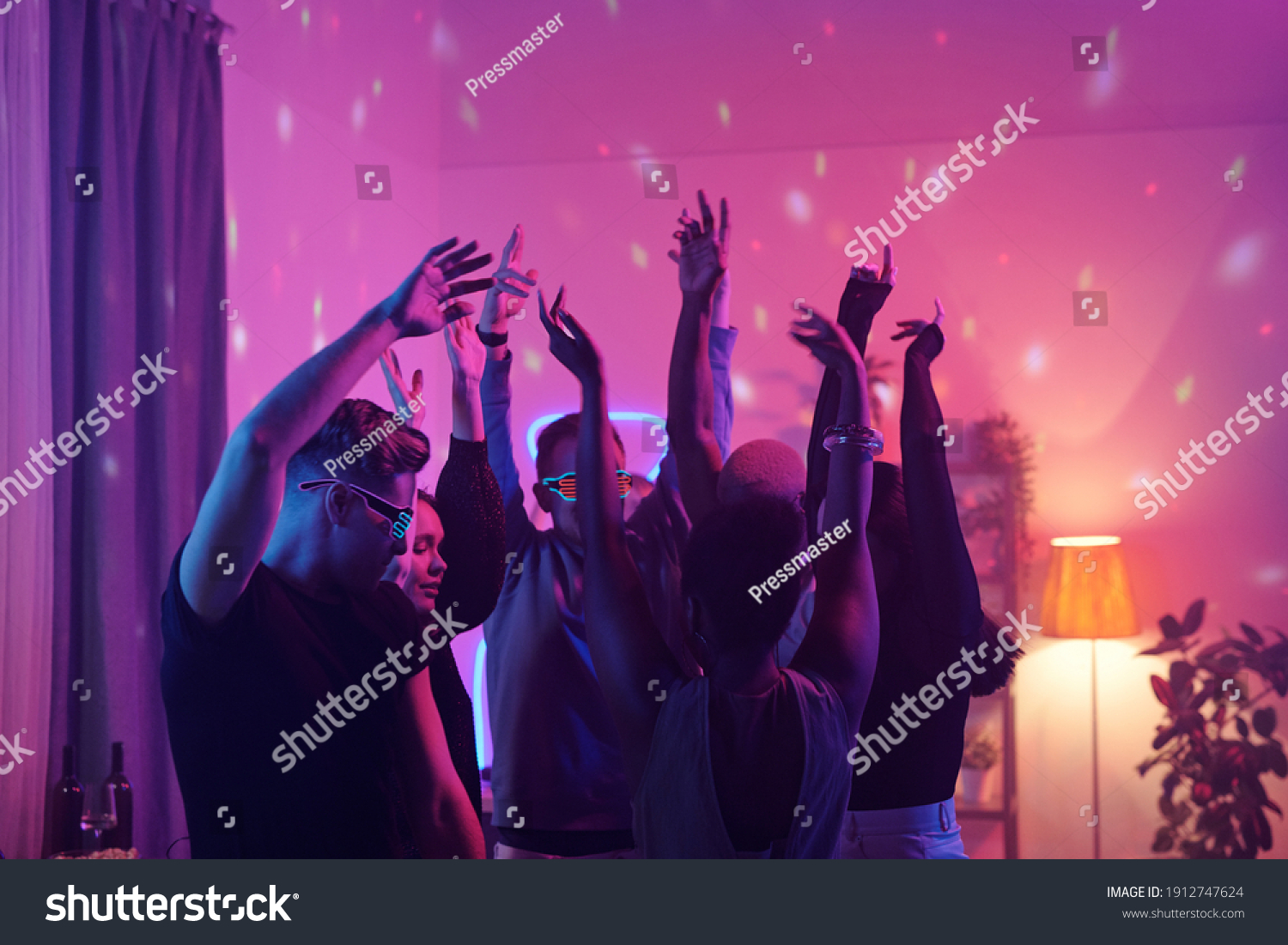 Young intercultural friends in smart casualwear raising arms while dancing together at home party in living-room illuminated with pink lighting #1912747624