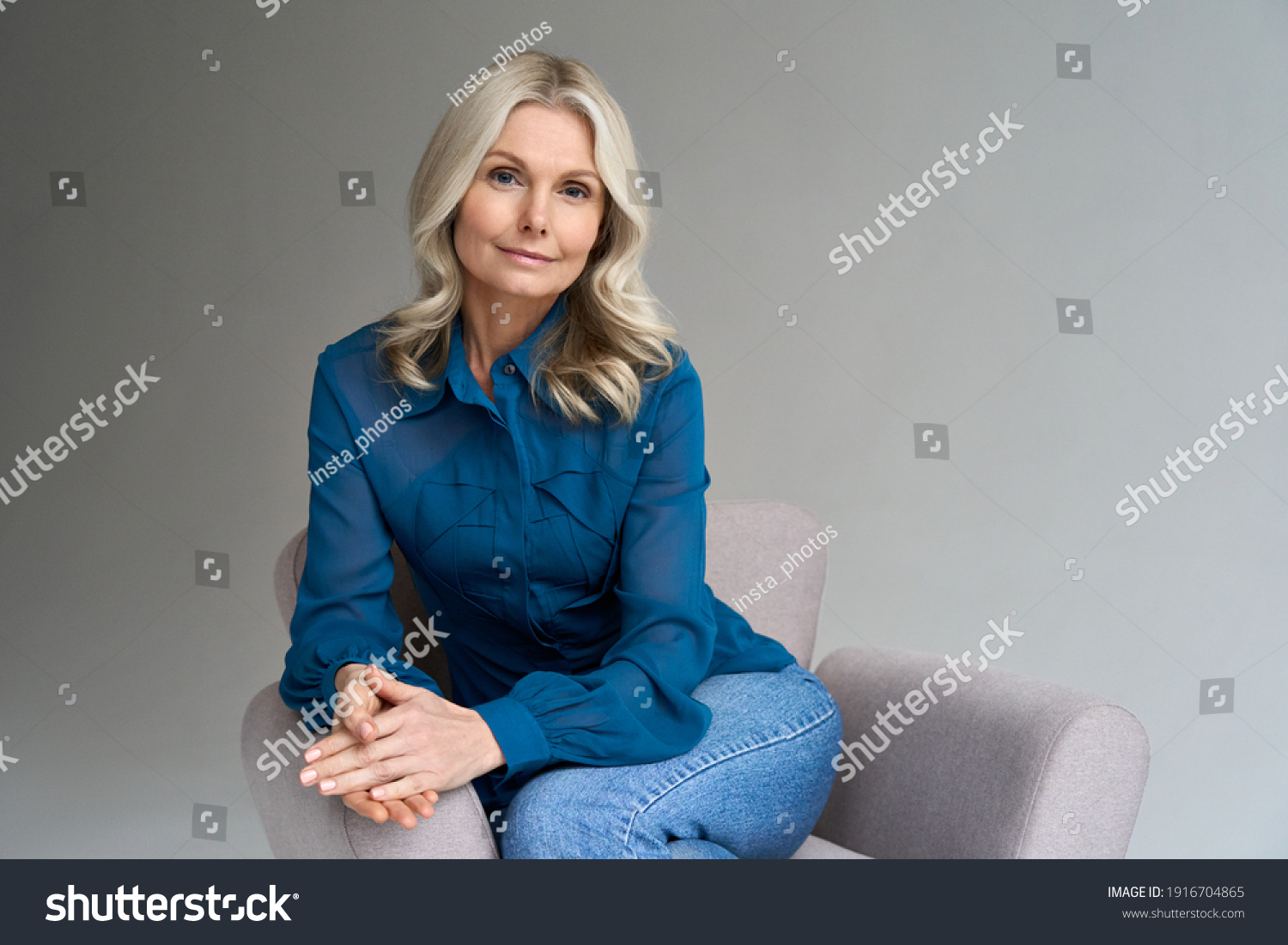 Middle aged female psychotherapist, counselor sitting in chair alone in office looking at camera. Sophisticated elegant mature 50s woman of mid age with blond hair posing indoors, portrait. #1916704865