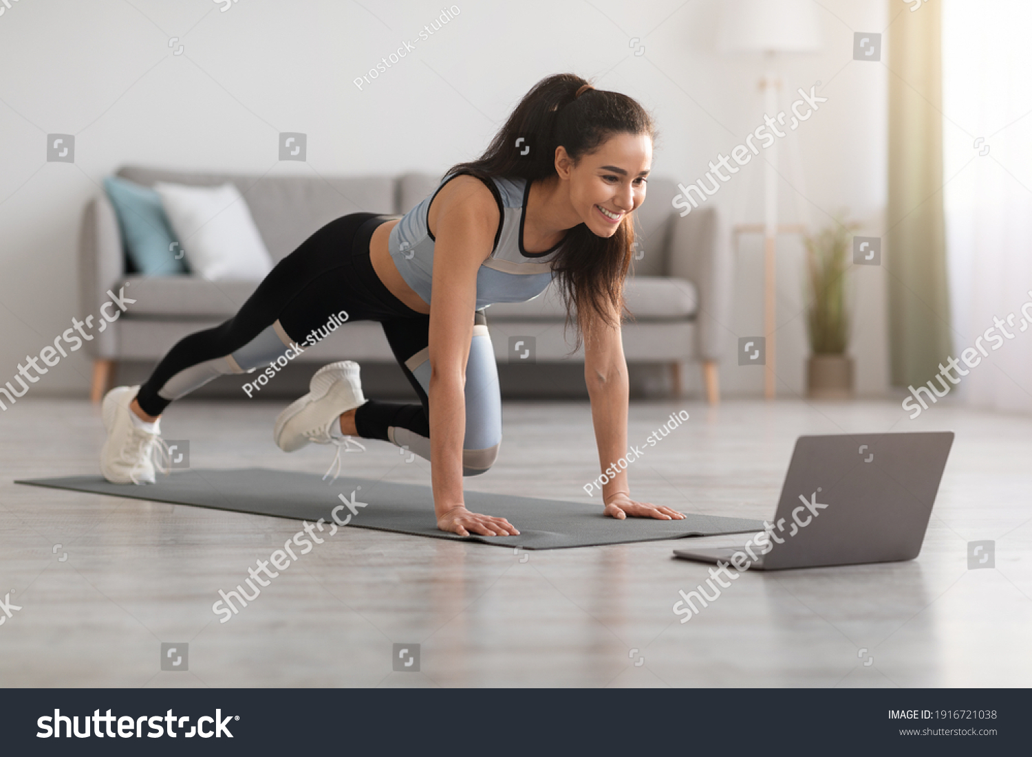 Cheerful active woman doing fitness at home, using laptop, watching online lessons. Positive well-fit lady in sportswear exercising during covid-19 pandemic, watching fitness videos on Internet #1916721038