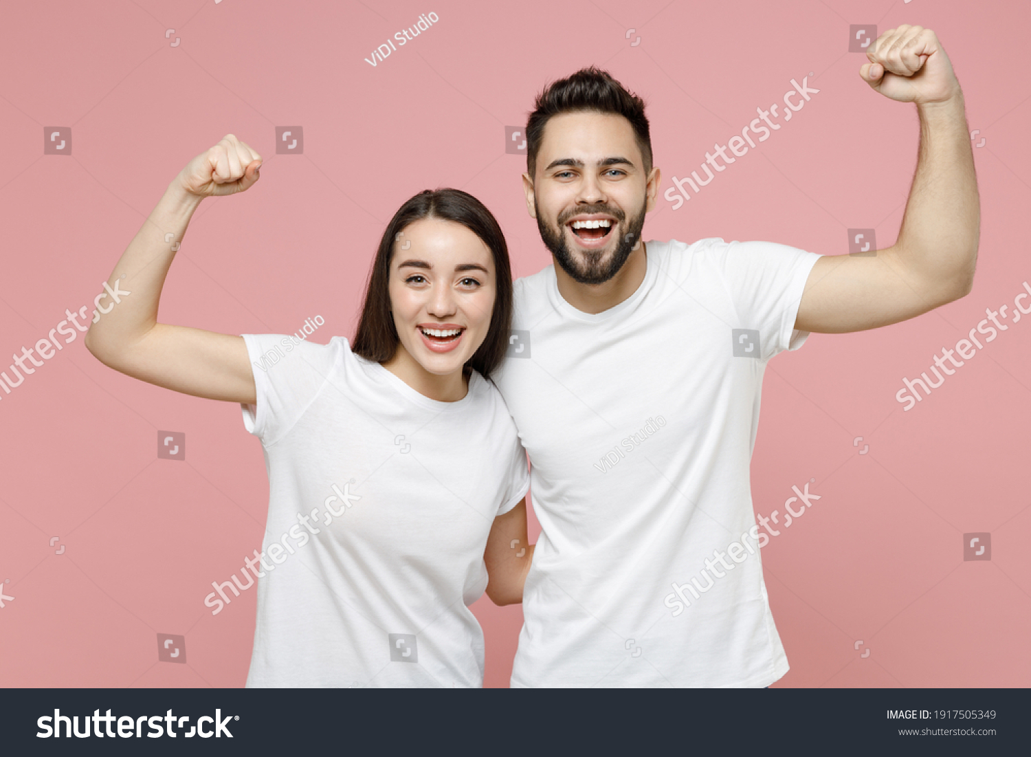 Young cheerful strong sporty fitness couple two friends man woman 20s in white basic blank print design t-shirts showing biceps muscles on hand isolated on pastel pink color background studio portrait #1917505349