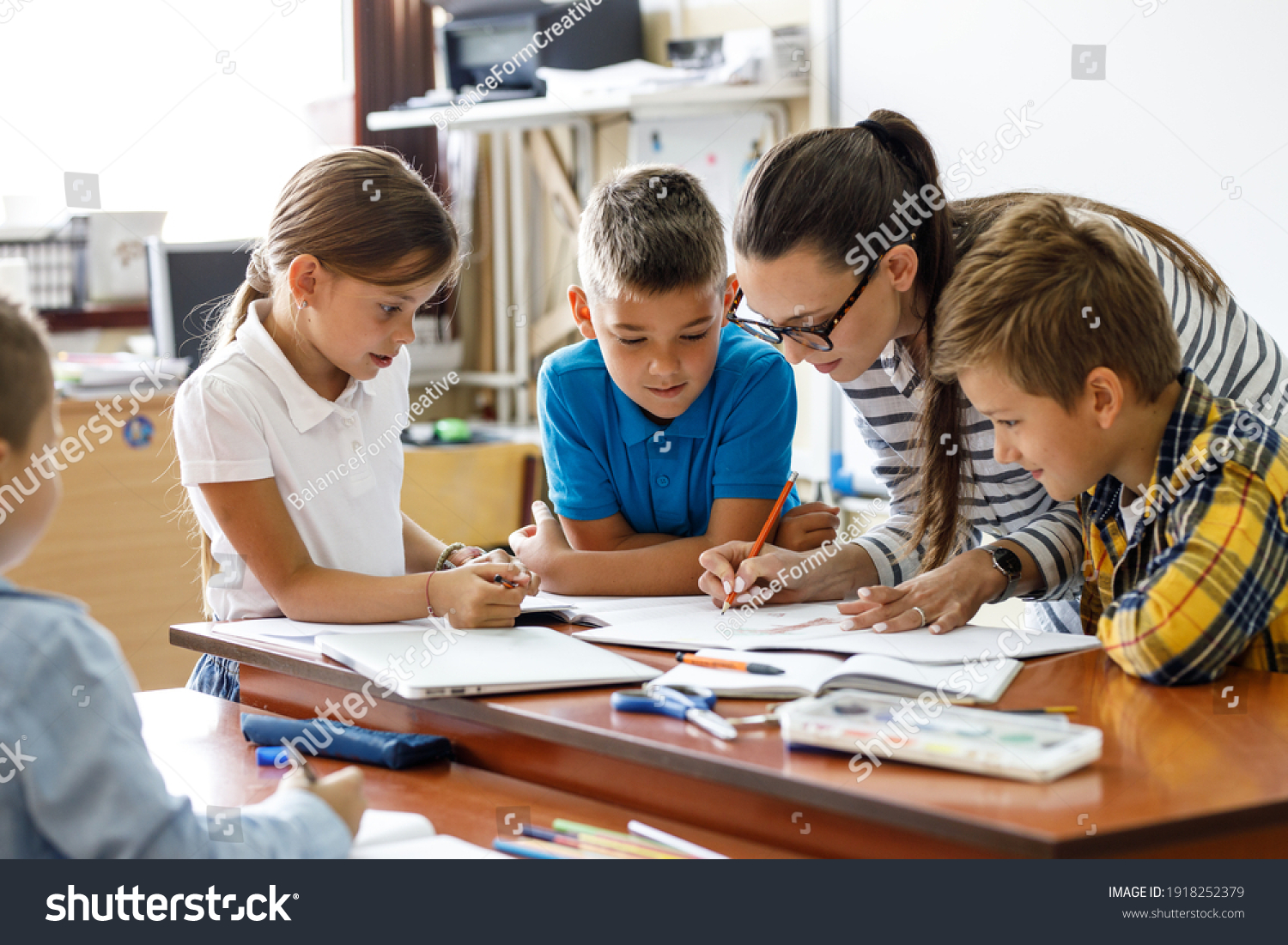 Female teacher helps school kids to finish they lesson.They sitting all together at one desk.	
 #1918252379