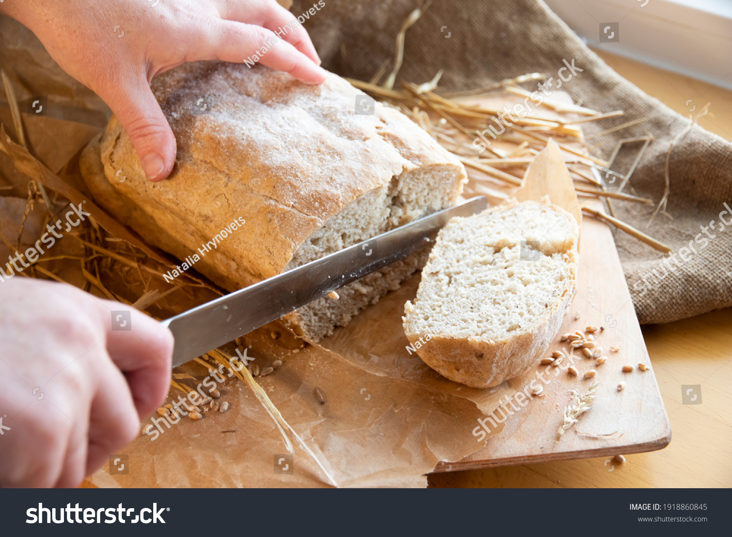 Woman slices fresh bread. hands with a knife. Freshly baked rye bread on a wooden board. Burlap, craft paper and straw on the background. Organic bread baked at home from wheat flour. Sliced bread. #1918860845