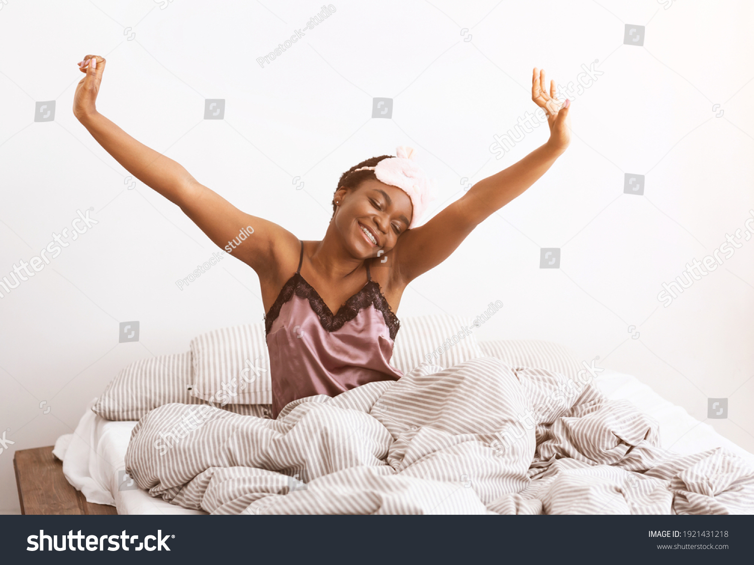 Happy and relaxed after good night sleep. Smiling millennial african american cute woman in mask sits on bed with blanket, stretching after waking up, in bedroom interior, copy space, lens flare #1921431218