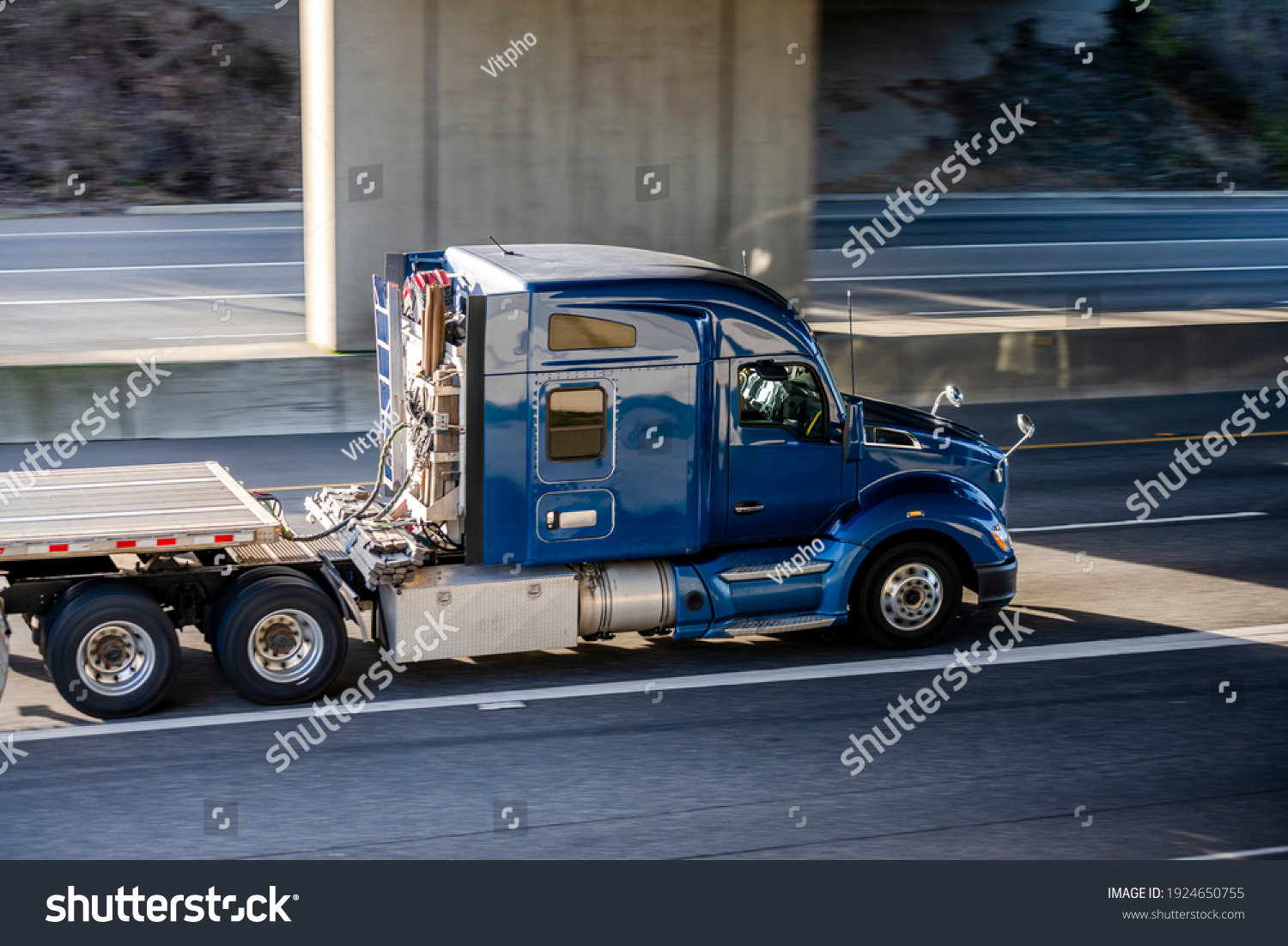 Big rig shiny blue semi truck tractor with long cab and auxiliary equipment on the back wall transporting flat bed semi trailer driving on the highway road under the concrete bridge #1924650755