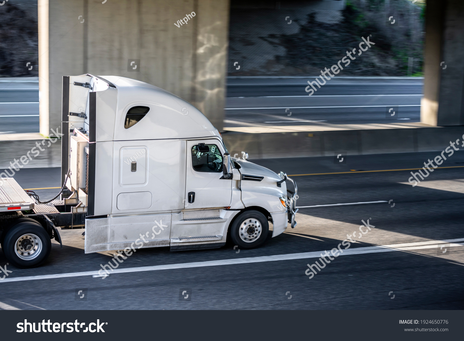 Big rig shiny white semi truck tractor with long cab and auxiliary equipment on the back wall transporting flat bed semi trailer driving on the highway road under the concrete bridge #1924650776