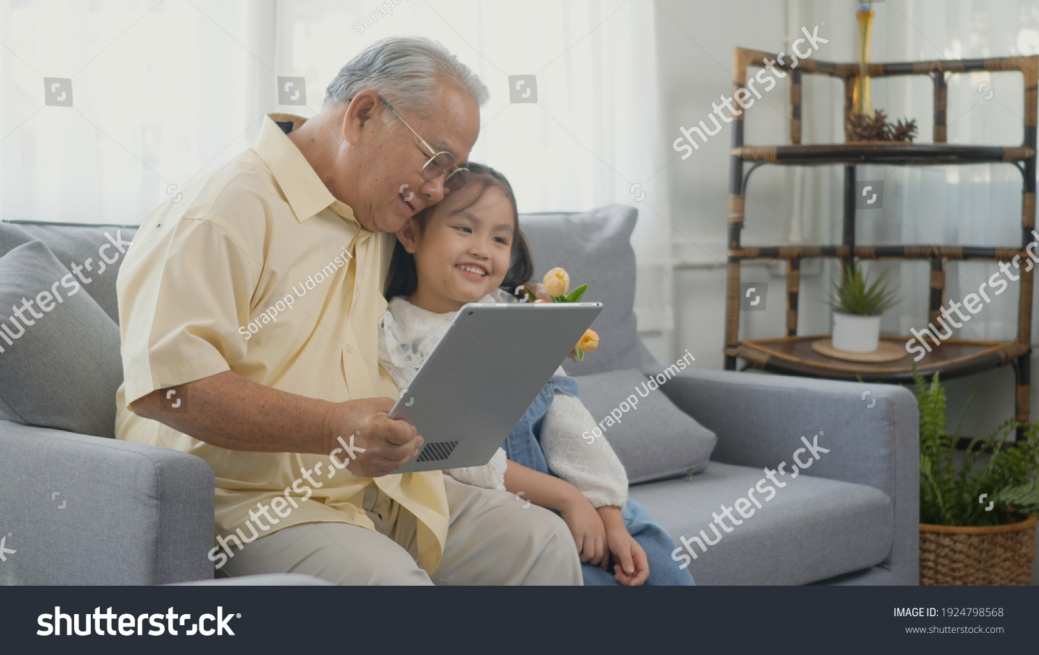 Asian senior old man looking to tablet computer and granddaughter come visitor at home, Grandfather reading news on digital tablet with his kid on sofa in living room #1924798568