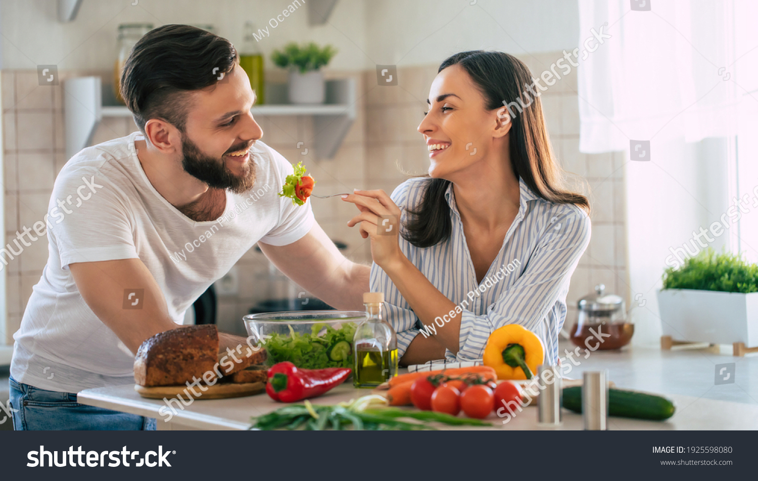 Excited smiling young couple in love making a super healthy vegan salad with many vegetables in the kitchen and man testing it from a girl's hands #1925598080