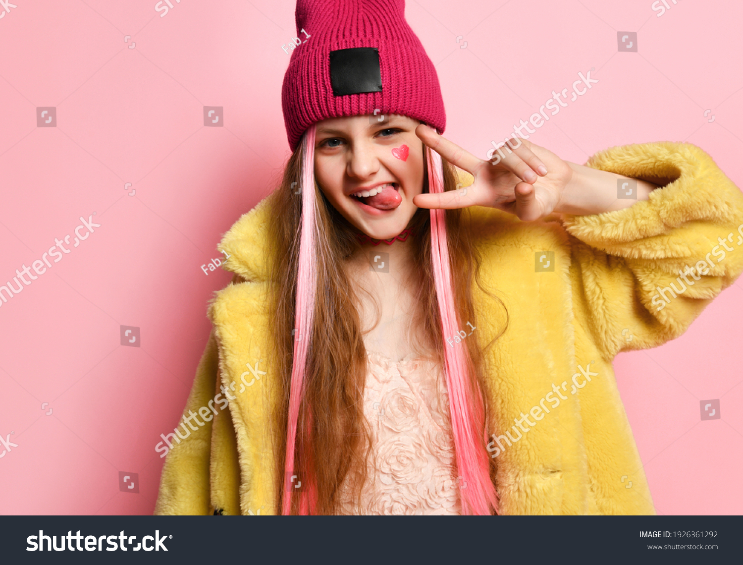 Close up portrait of an active and cheerful teenage girl who is showing V-sign with her fingers and tongue. Little rebel in a pink hat, yellow fur coat posing on a pink background. Adolescence concept #1926361292