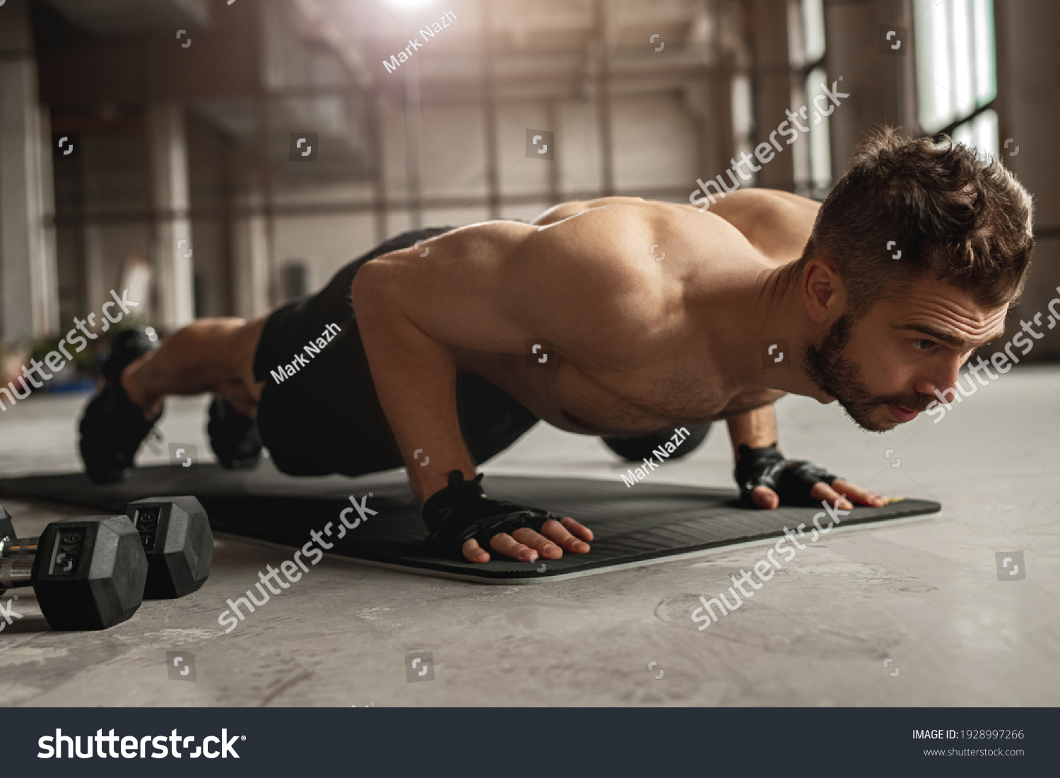 Muscular man doing push up exercise on mat near dumbbells during intense fitness workout in grungy gym #1928997266