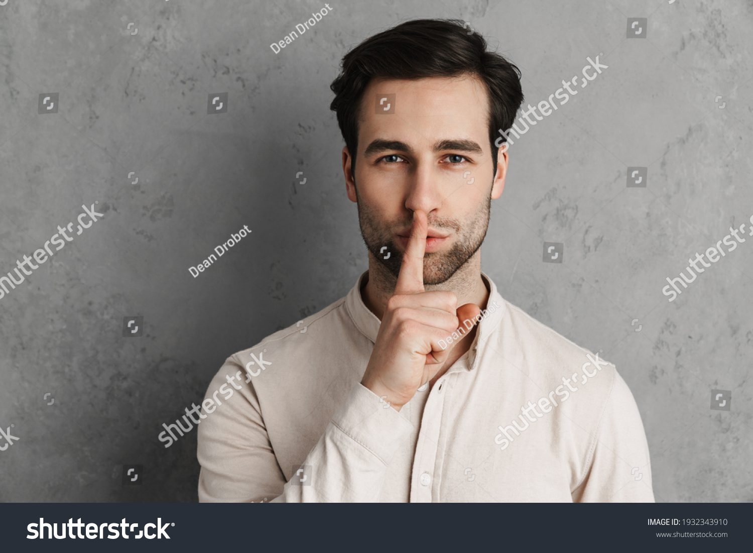Smiling young man asking for silence gesturing with his finger isolated over gray background #1932343910