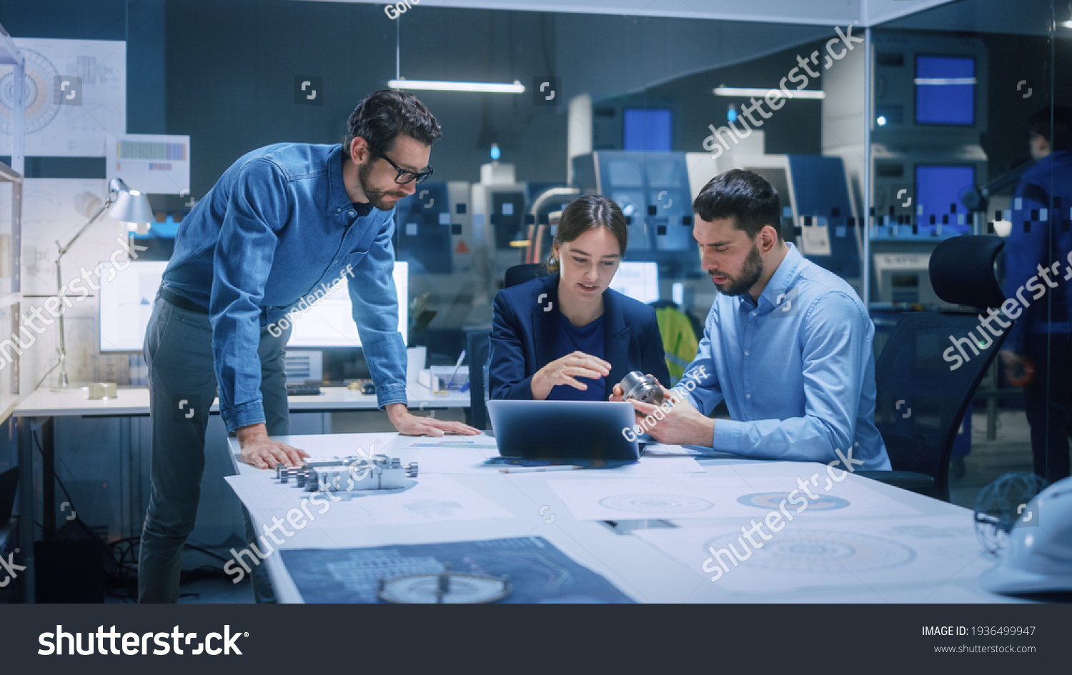 Factory Office Meeting Room: Team of Engineers Gather Around Conference Table, They Discuss Project Blueprints, Inspect Mechanism, Find Solutions, Use Laptop. Industrial Technology Factory #1936499947