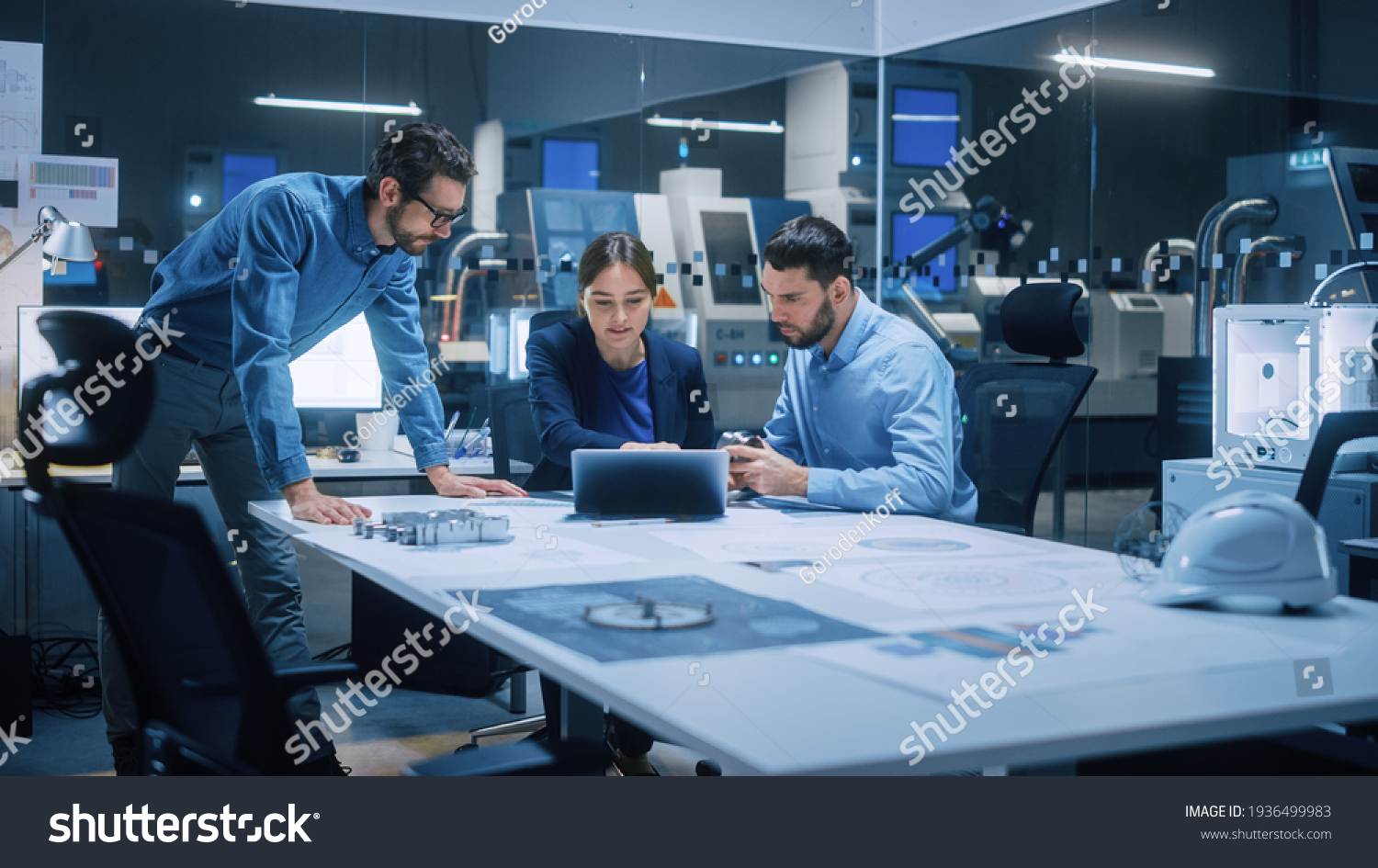 Factory Office Meeting Room: Team of Engineers Gather Around Conference Table, They Discuss Project Blueprints, Inspect Mechanism, Find Solutions, Use Laptop. Industrial Technology Factory #1936499983