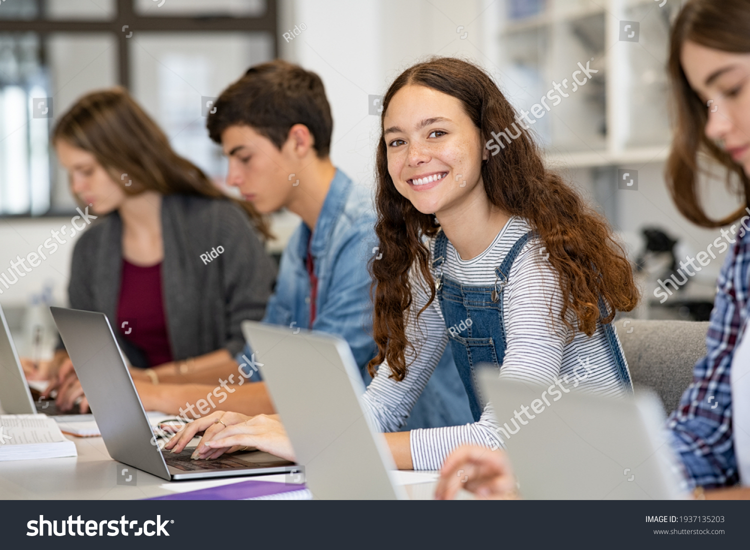 Happy young woman working on laptop and looking at camera in classroom. Portrait of smiling university student in library use computer for a research. Satisfied college student looking at camera. #1937135203