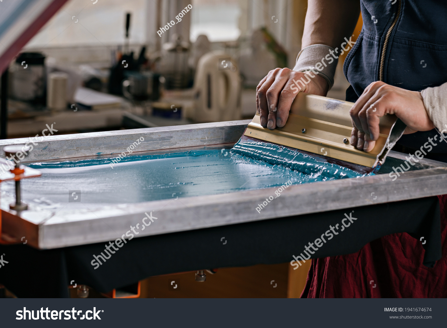 selective focus photo of male hands with squeegee. serigraphy production. printing images on t-shirts by silkscreen method in a design studio #1941674674