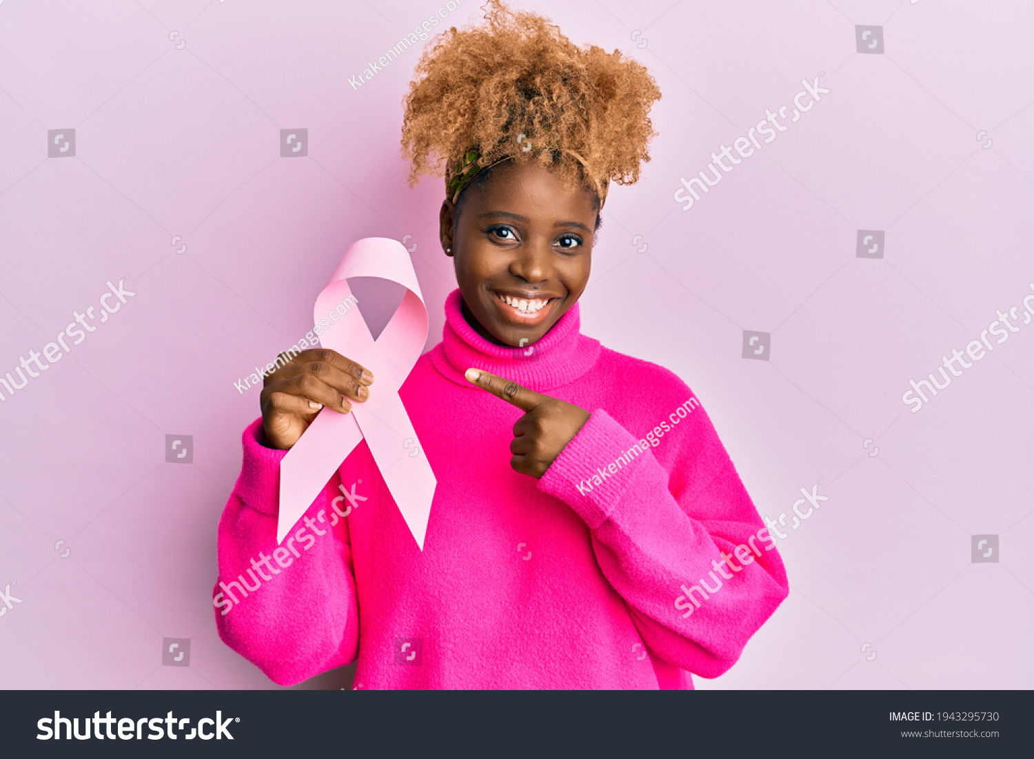 Young african woman with afro hair holding pink cancer ribbon smiling happy pointing with hand and finger  #1943295730