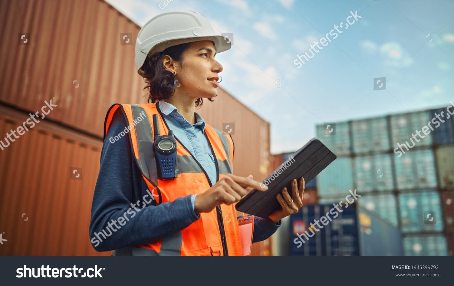 Smiling Portrait of a Beautiful Latin Female Industrial Engineer in White Hard Hat, High-Visibility Vest Working on Tablet Computer. Inspector or Safety Supervisor in Container Terminal. #1945399792