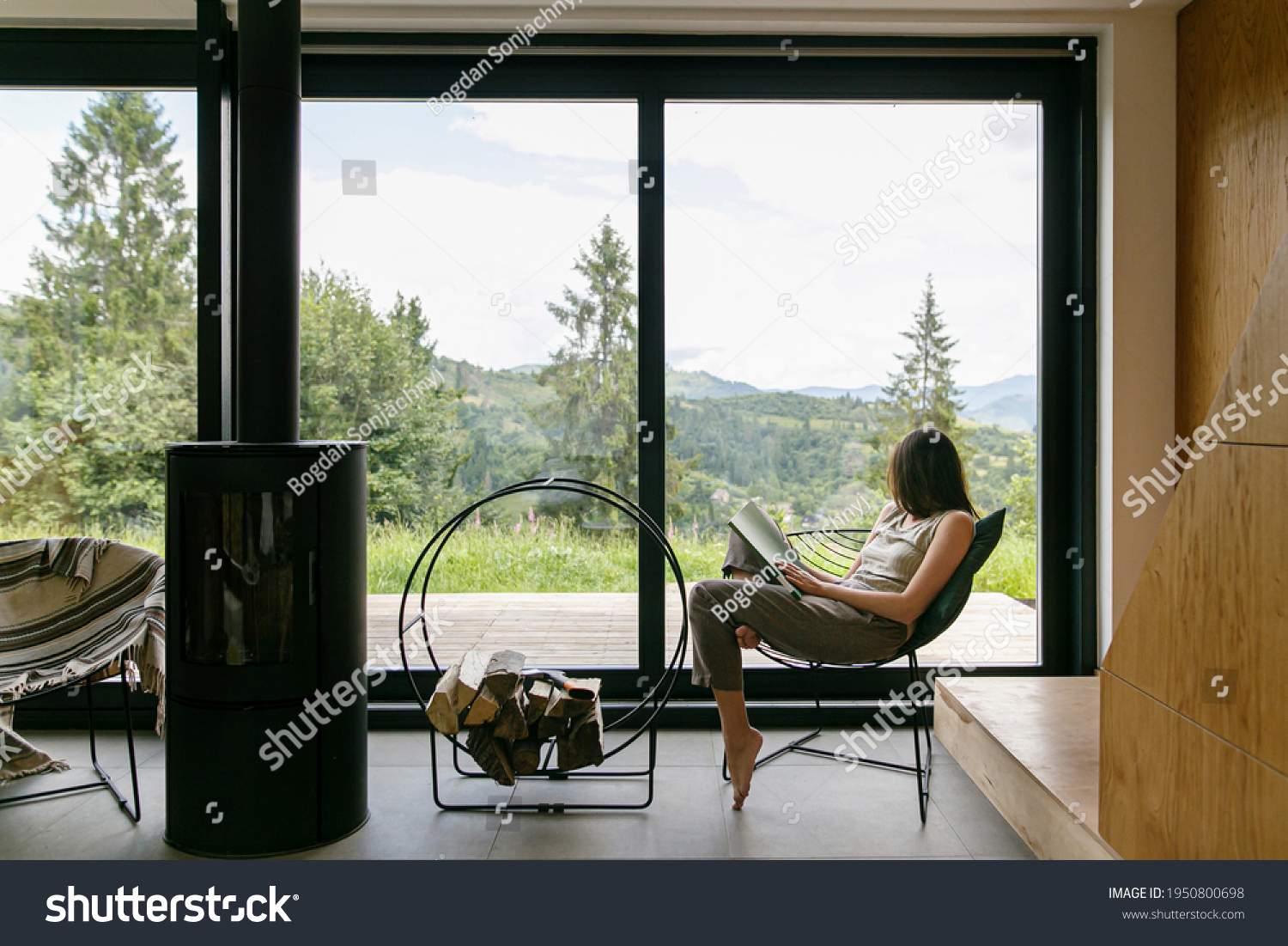 Beautiful stylish woman reading book on chair at fireplace with firewood on background of mountain hills. Young female in casual clothes relaxing in modern chalet with amazing view from window #1950800698