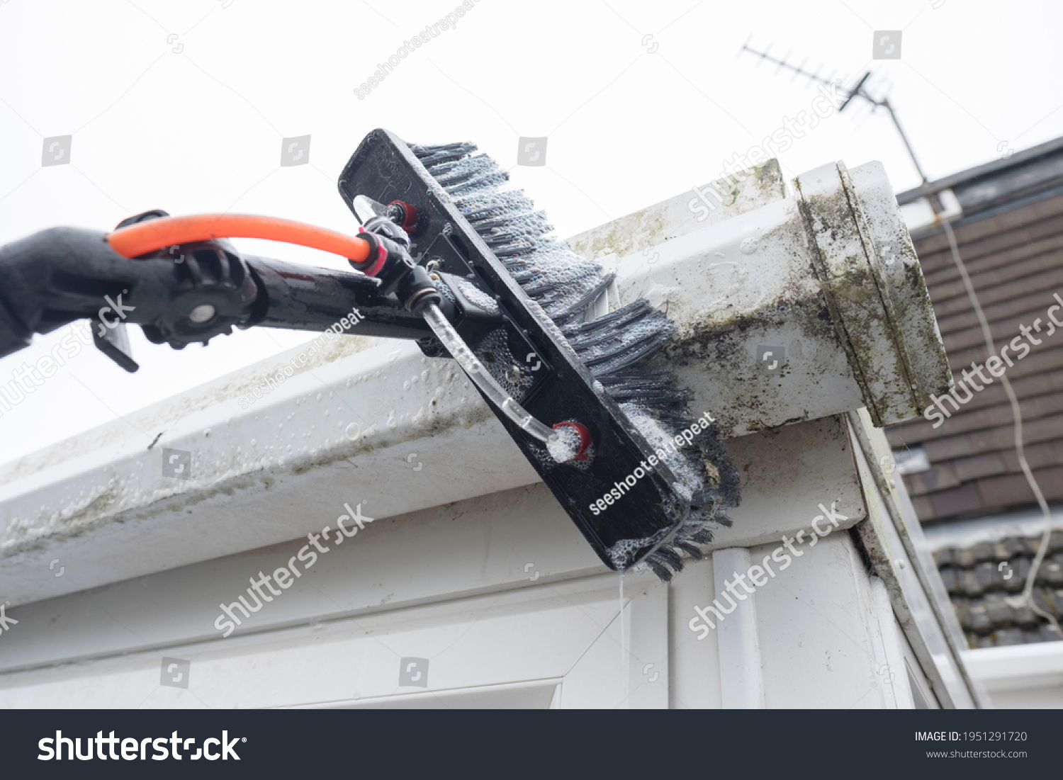 A brush cleaning dirty clogged white plastic pvc gutters and drain pipes with mossy green mould plastic fascias.  Blocked drains and guttering need window cleaners and regular yard work maintenance  #1951291720