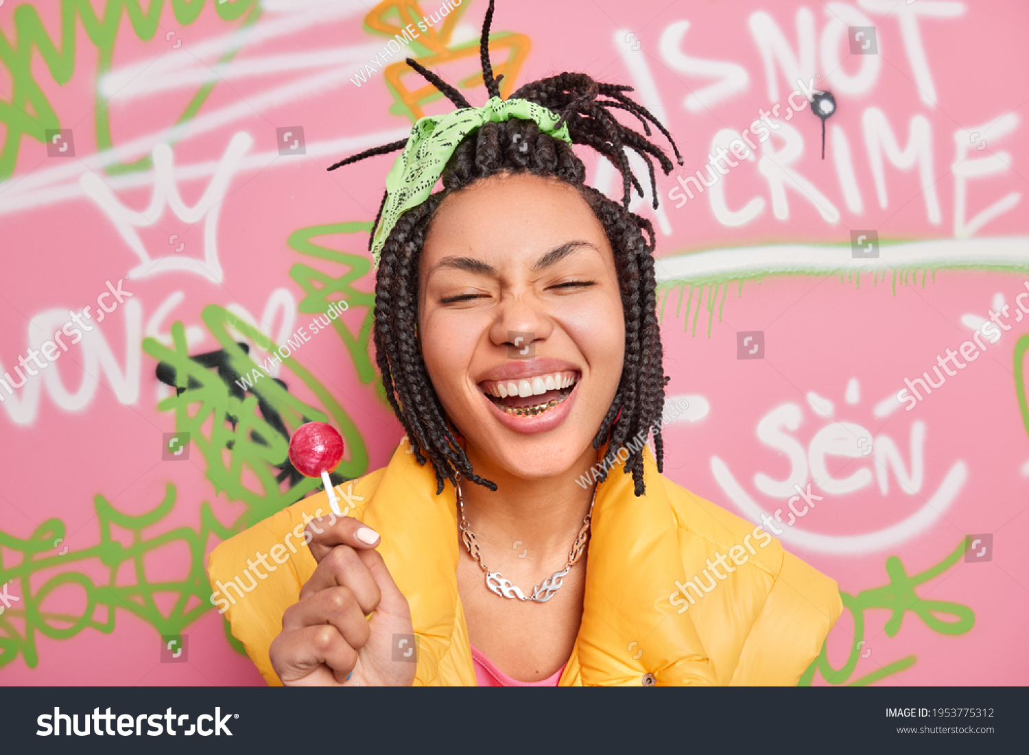 Trendy cheerful hipster girl smiles broadly holds lollipop has fun with teenagers of same age wears yellow vest poses against colorful graffiti wall. Gen z generation. Youth lifestyle concept #1953775312