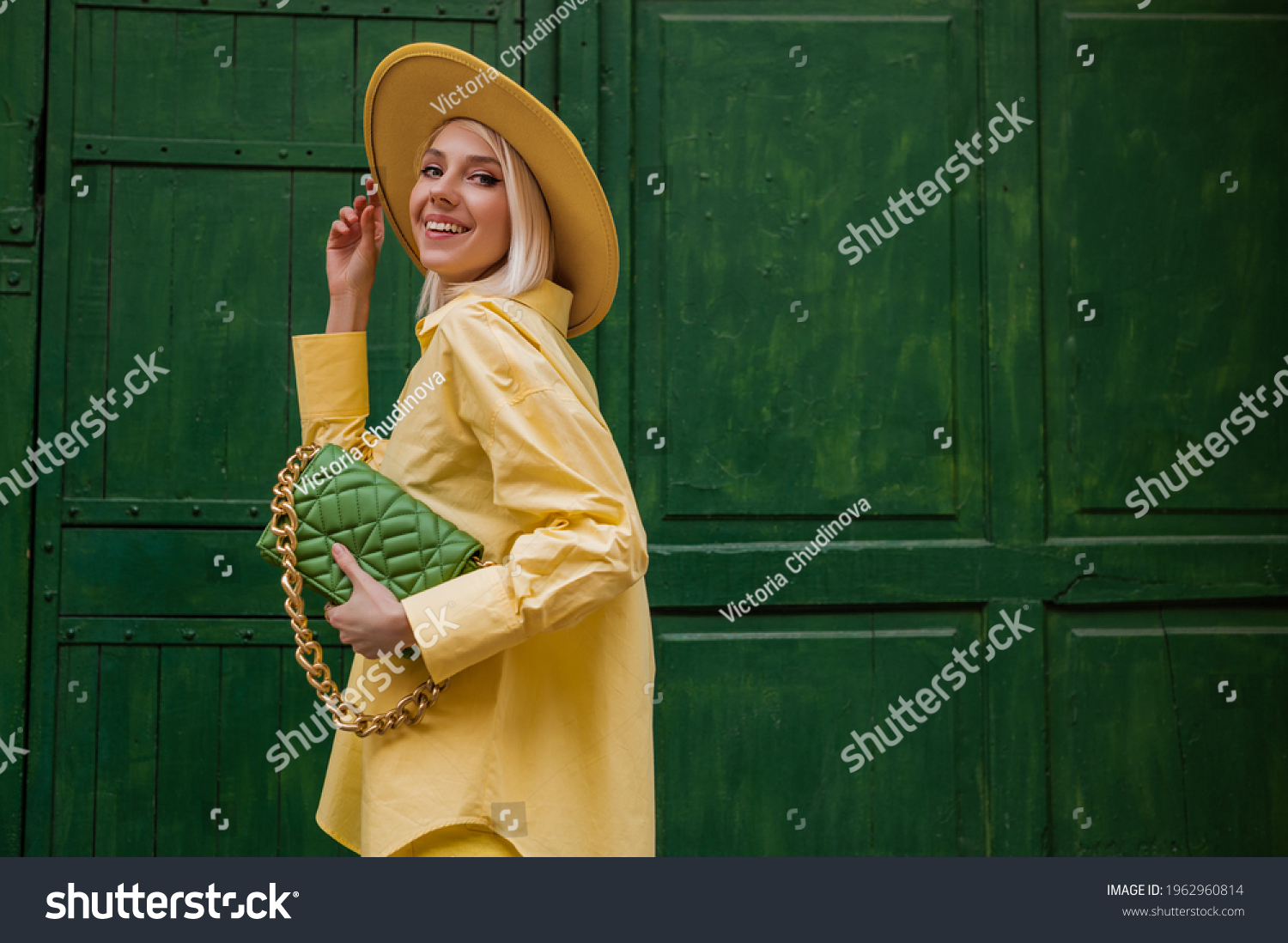 Joyful smiling woman wearing yellow hat, classic shirt, holding quilted faux leather green bag with chain, posing on green background in street. Copy, empty space for text #1962960814