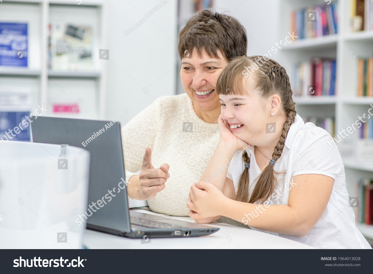 Happy teacher and smiling girl with down syndrome use a laptop at library. Education for disabled children concept #1964013028