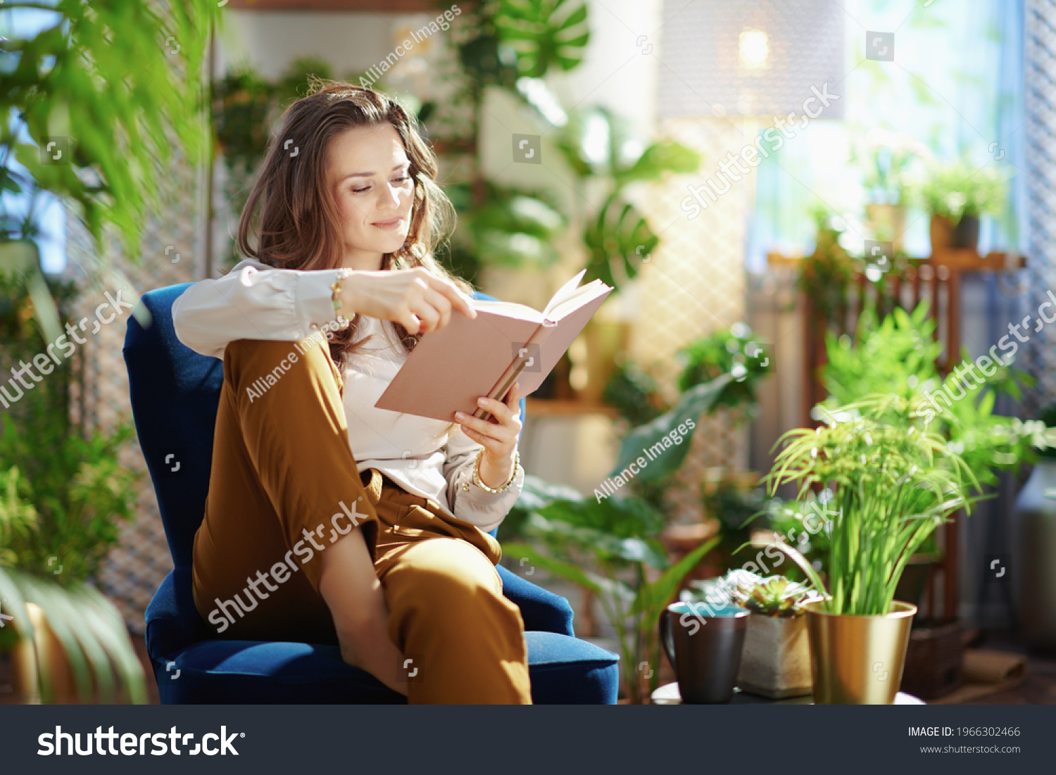 Green Home. relaxed trendy middle aged woman with long wavy hair with book in green pants and grey blouse in the modern living room in sunny day. #1966302466