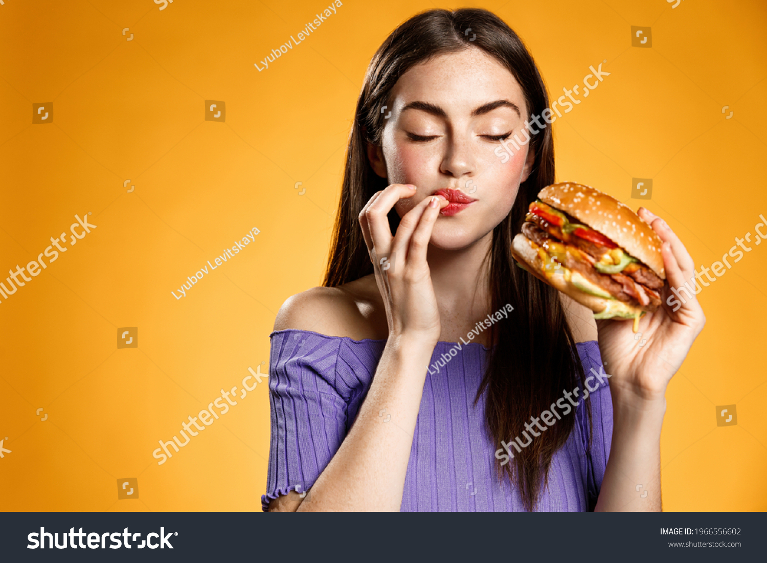 Woman eating cheeseburger with satisfaction. Girl enjoys tasty hamburger takeaway, licking fingers delicious bite of burger, order fastfood delivery while hungry, standing over orange background #1966556602