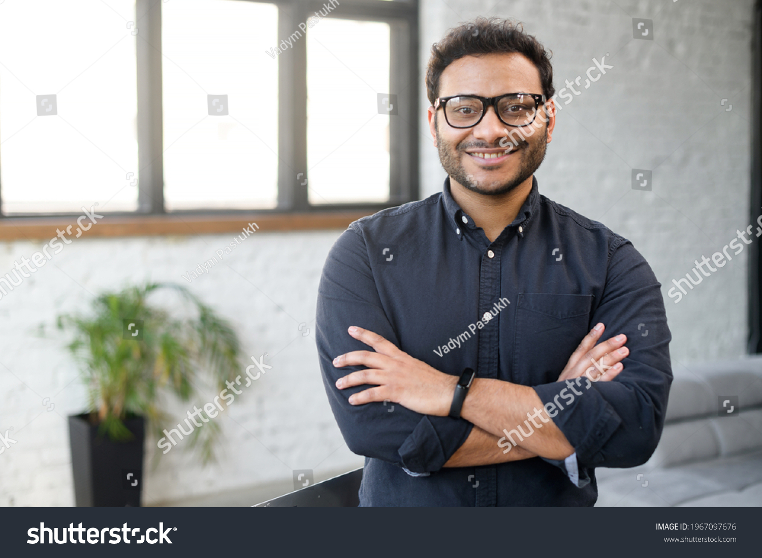 Headshot of skilled hindu male employee standing with arms crossed in modern office, successful confident mixed-race man wearing eyeglasses and smart casual, business portrait of indian entrepreneur #1967097676