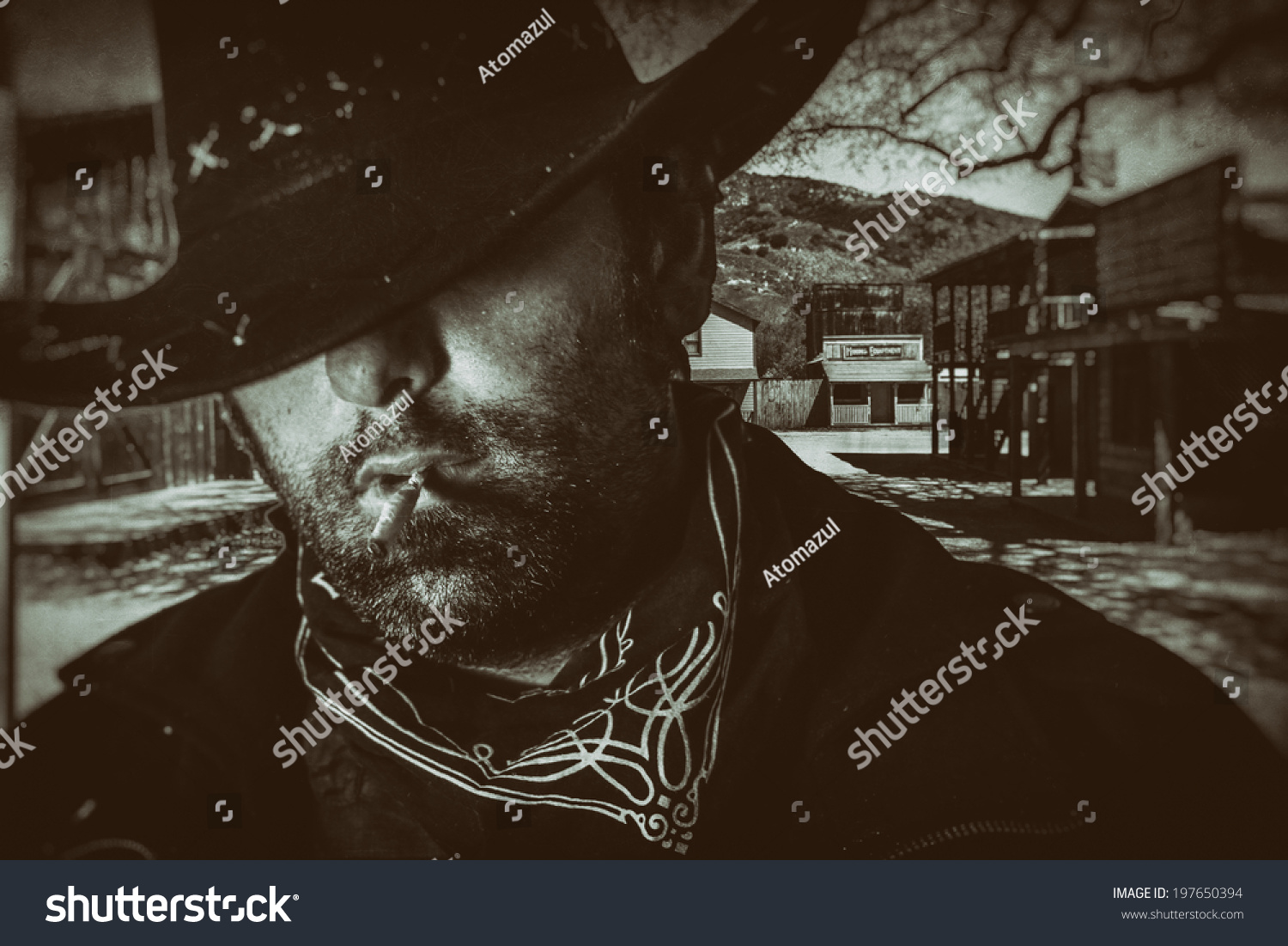 Old West Cowboy Western Town. An old west cowboy in a hat smoking a hand rolled cigarette with an old western town setting in the background, edited in vintage film style. #197650394