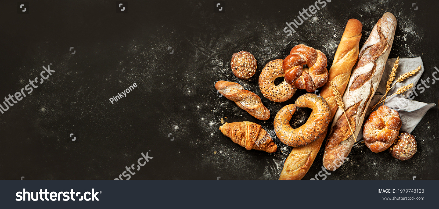 Bakery - various kinds of breadstuff. Bread rolls, baguette, bagel, sweet bun and croissant captured from above (top view, flat lay). Black background, free copy space. Horizontal banner layout. #1979748128