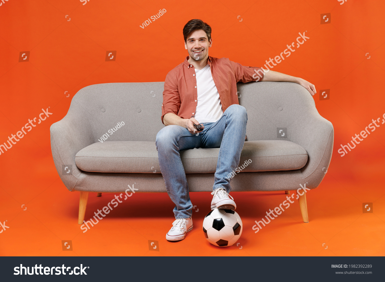 Young man football fan in shirt support favorite team with soccer ball sit on sofa at home watch tv live stream switch channel isolated on orange background. People sport leisure lifestyle concept. #1982392289