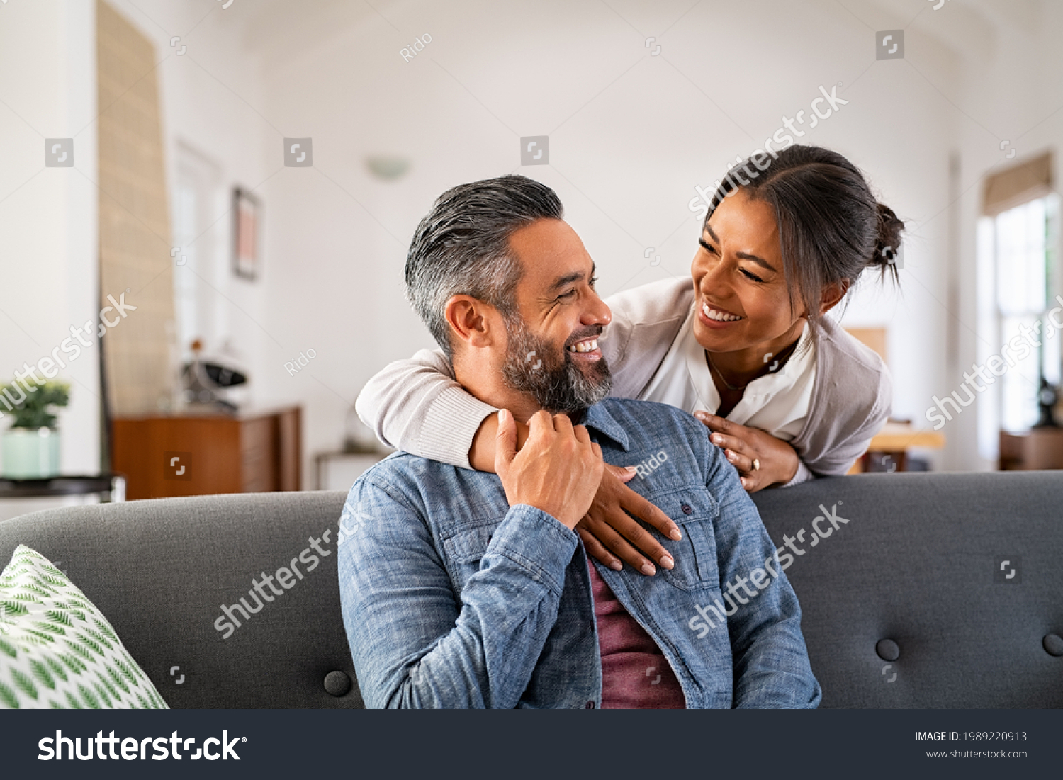 Smiling ethnic woman hugging her husband on the couch from behind in the living room. Middle eastern man having fun with his beautiful young wife on the couch. Mid adult indian man with latin woman. #1989220913