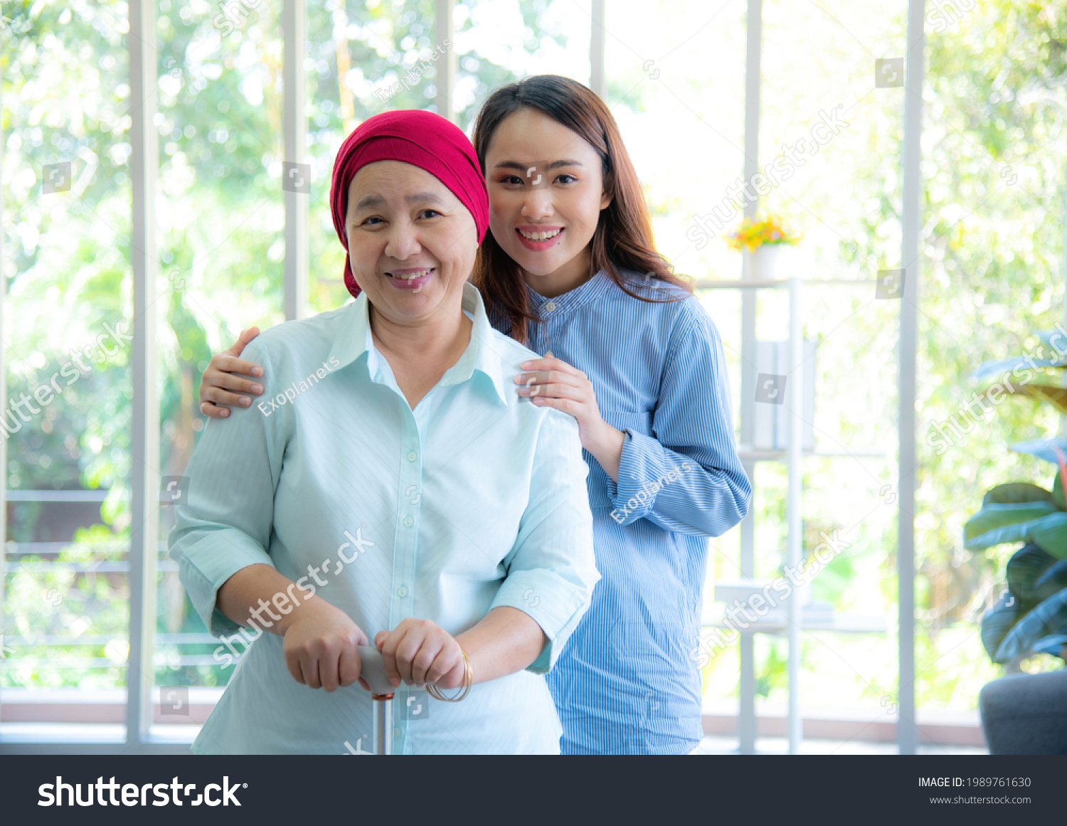 Asian elderly mother wearing red headscarf recover from cancer standing, holding walking stick and having her daughter by her side embrace, smile very happily. Cancer or leukemia survivor concept. #1989761630