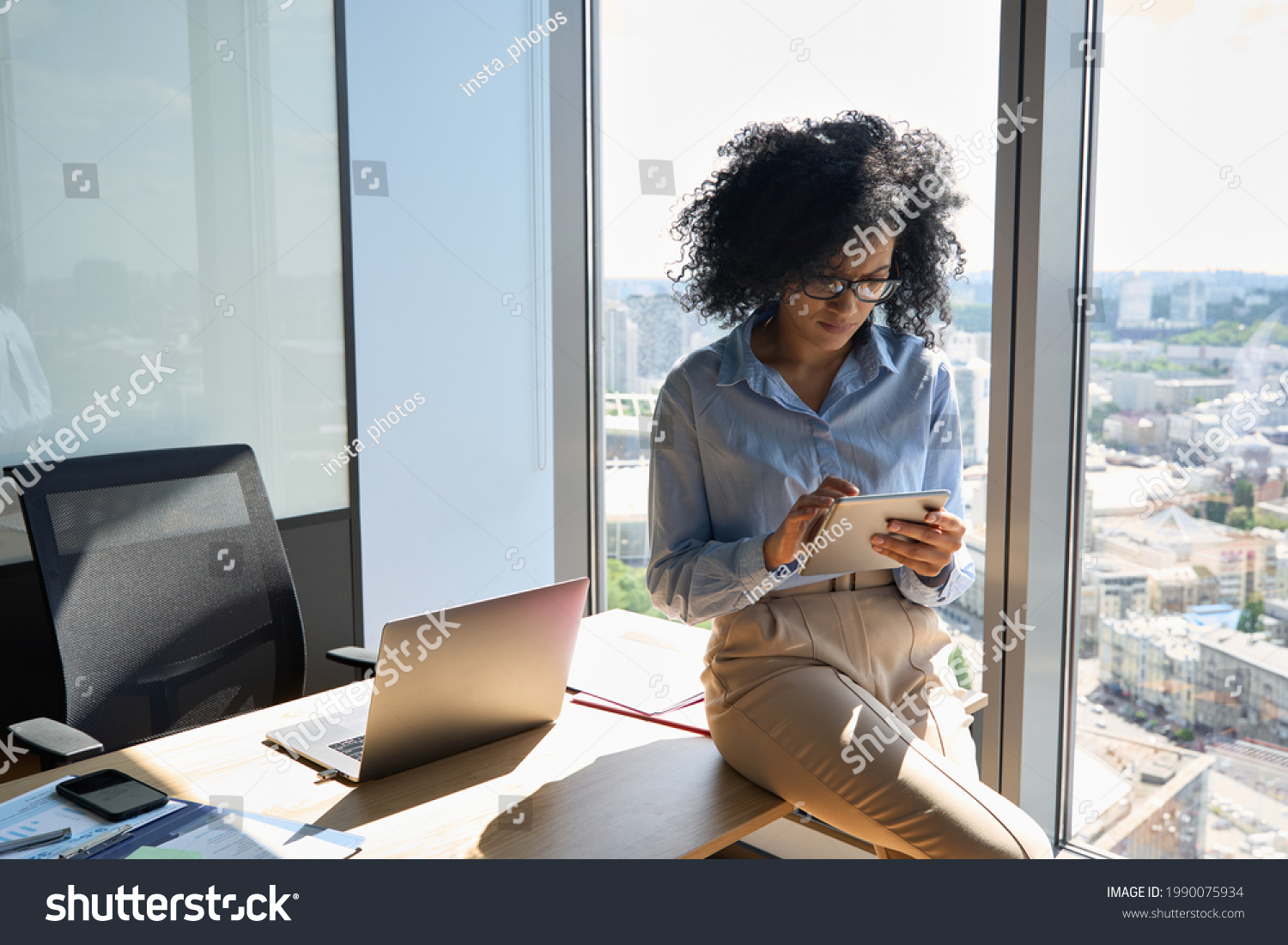 Young focused African American businesswoman sitting on desk holding using tablet computer device in modern office in high floor building with panoramic urban capital city view. #1990075934