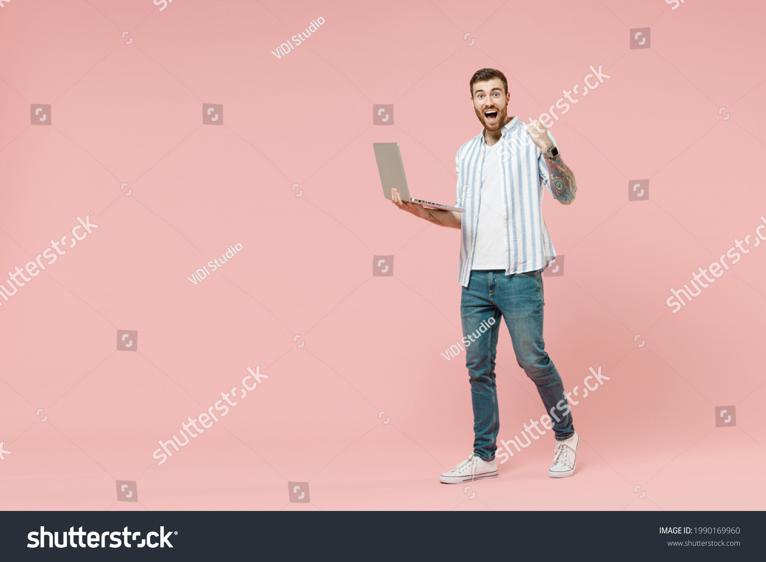 Full length young smiling happy fun unshaven man in blue striped shirt holding laptop pc computer chat online browsing internet do winner gesture clench fist isolated on pastel pink background studio. #1990169960