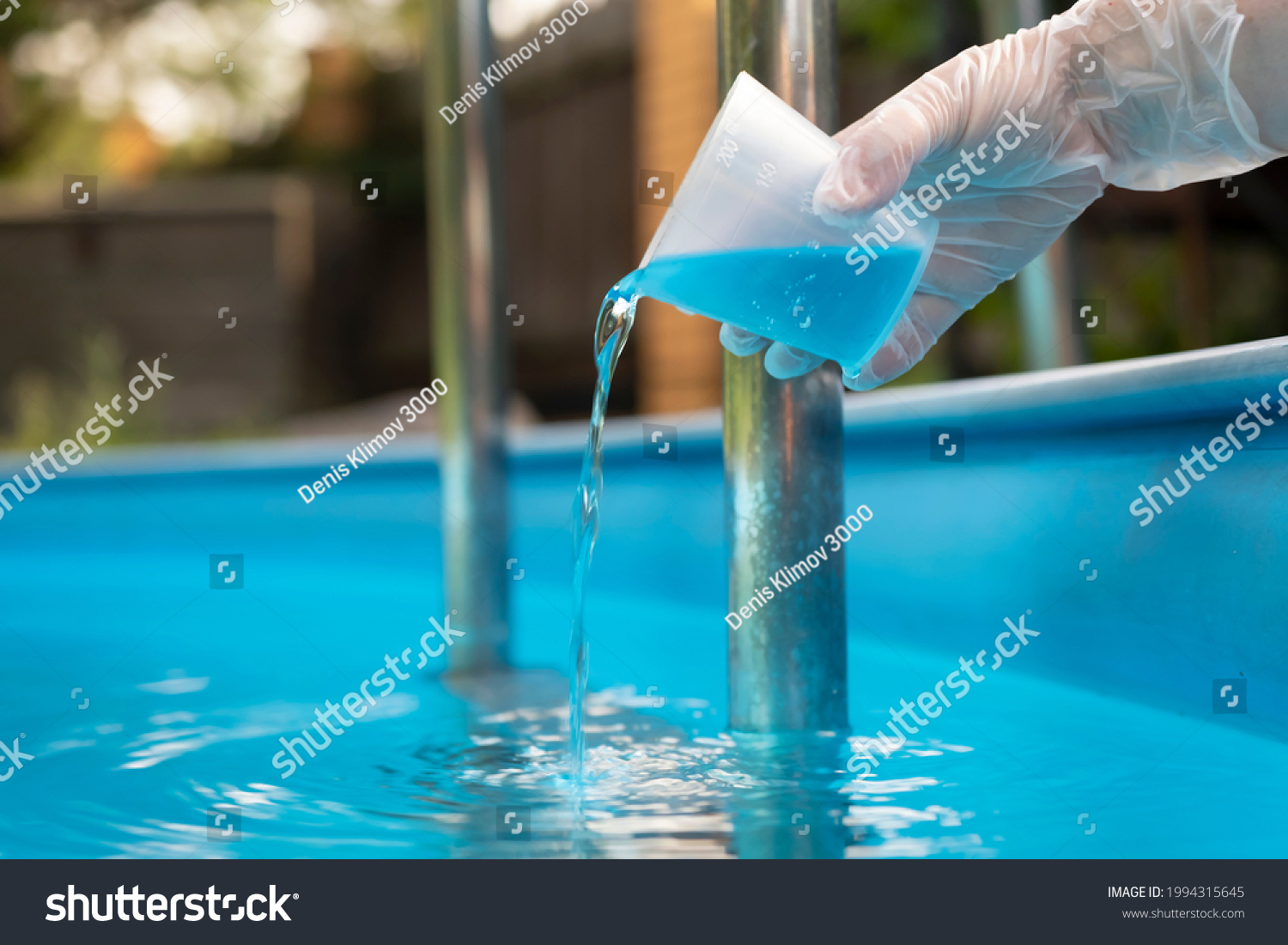 Hand holding and pouring algicide blue liquid from glass into water of swimming pool. Water purification and prevention of appearance of algae and microorganisms. #1994315645