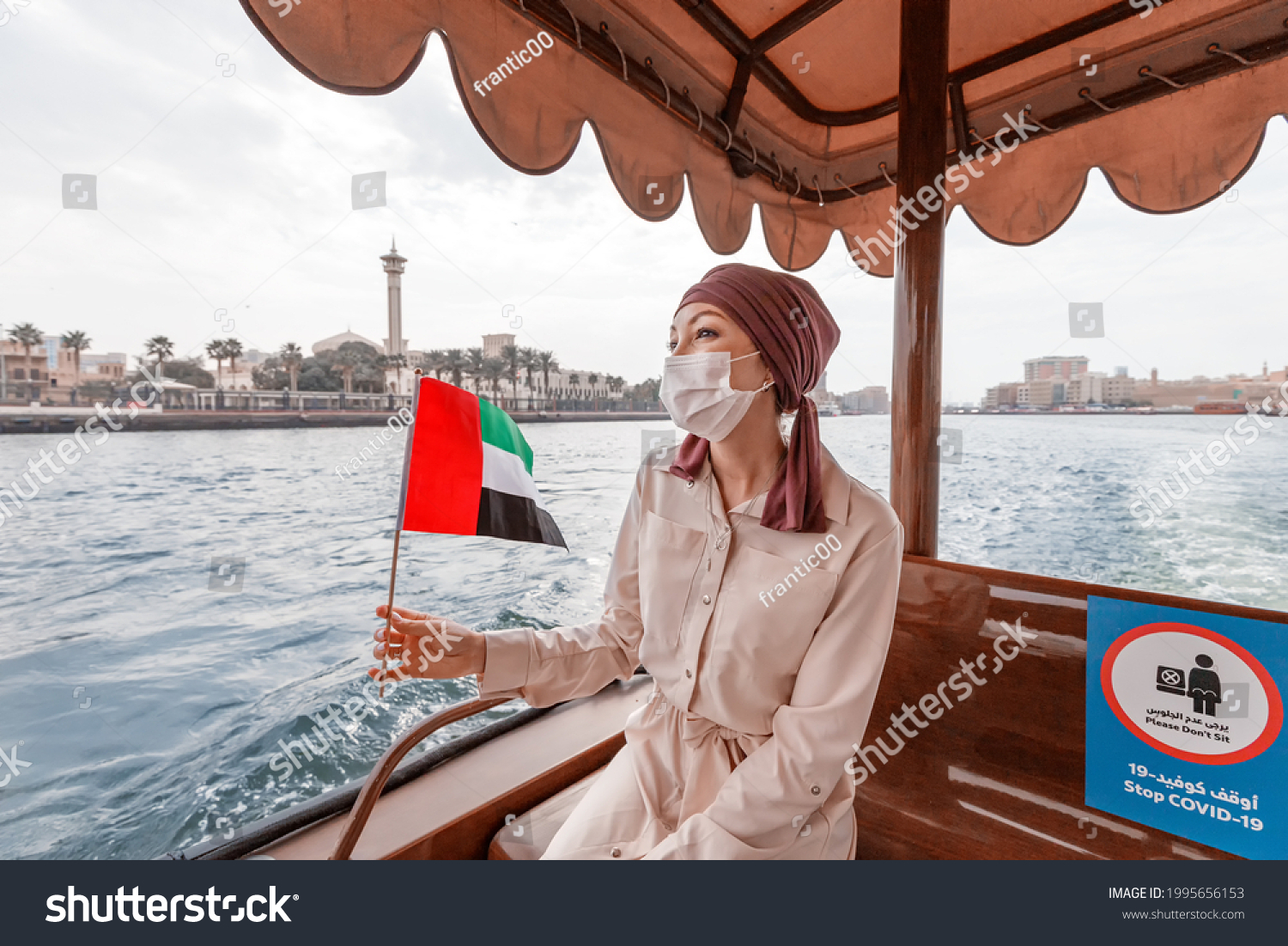 Happy woman wearing a medical mask with the flag of the UAE is traveling on the traditional boat ferry Abra Dhow on the Dubai Creek. Coronavirus and covid-19 are not a hindrance to travel #1995656153