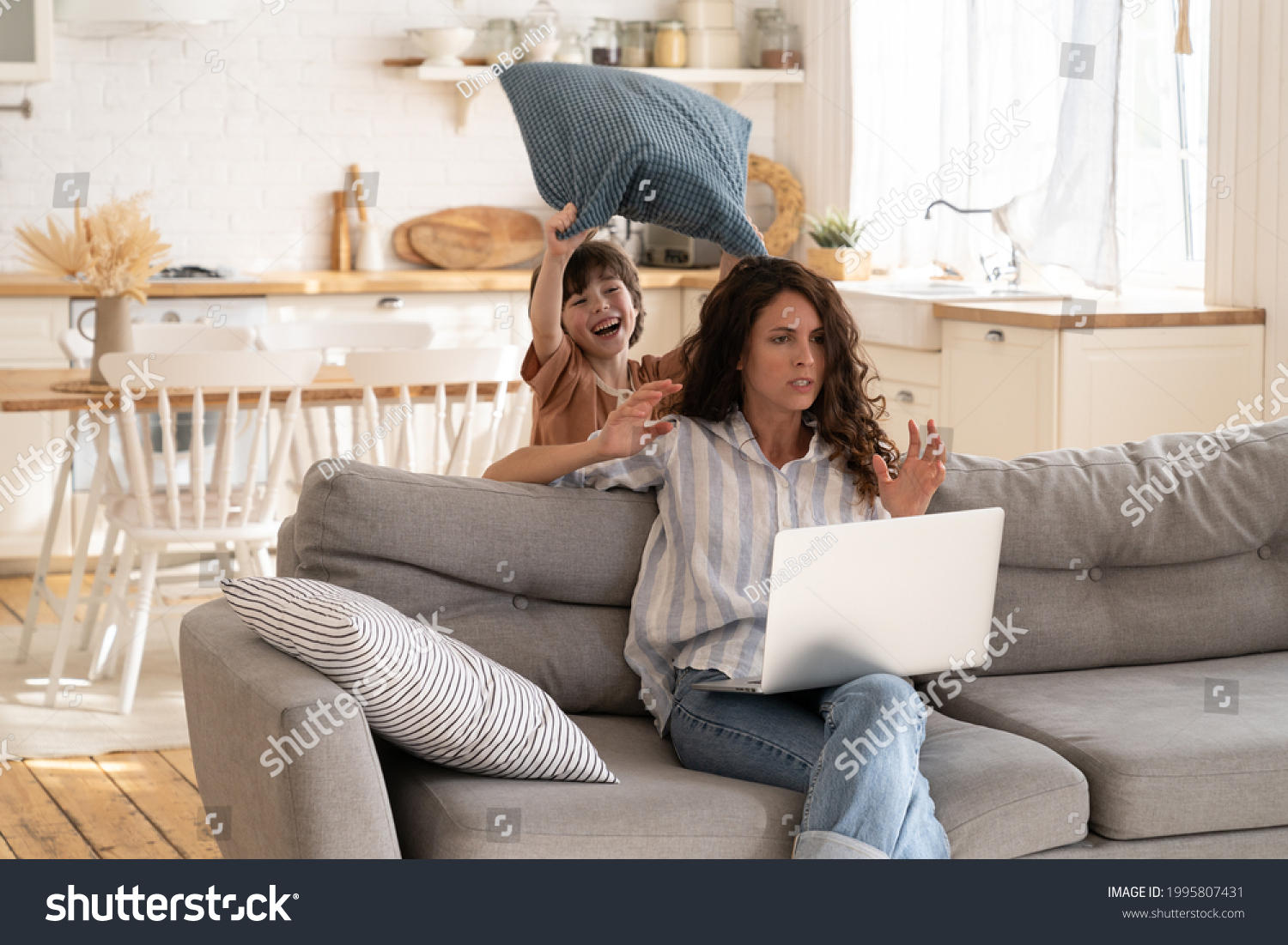 Furious young woman try to concentrate on work as small disobedient kid hit her with pillow. Angry mother freelancer or distance office worker on lockdown at home with hyperactive preschooler child #1995807431